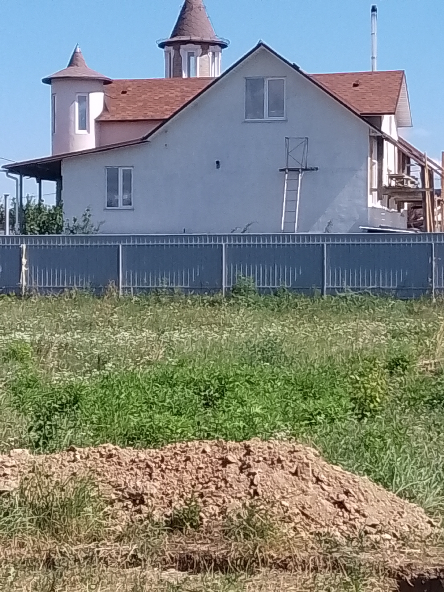 In Zalizne, Ukraine, a construction site is visible under a clear blue sky. A partially built house with a reddish-brown shingle roof and pale exterior walls dominates the scene. The house features several windows and two distinctive turret-like structures, one with a conical roof and another with a more rounded cap. A metal ladder leans against the side of the house, suggesting ongoing work. A grey metal fence runs along the property line, separating the construction site from an overgrown grassy area. In the foreground, a pile of reddish-brown soil is visible amidst the tall grass. The overall impression is one of active construction in a rural or semi-rural setting.