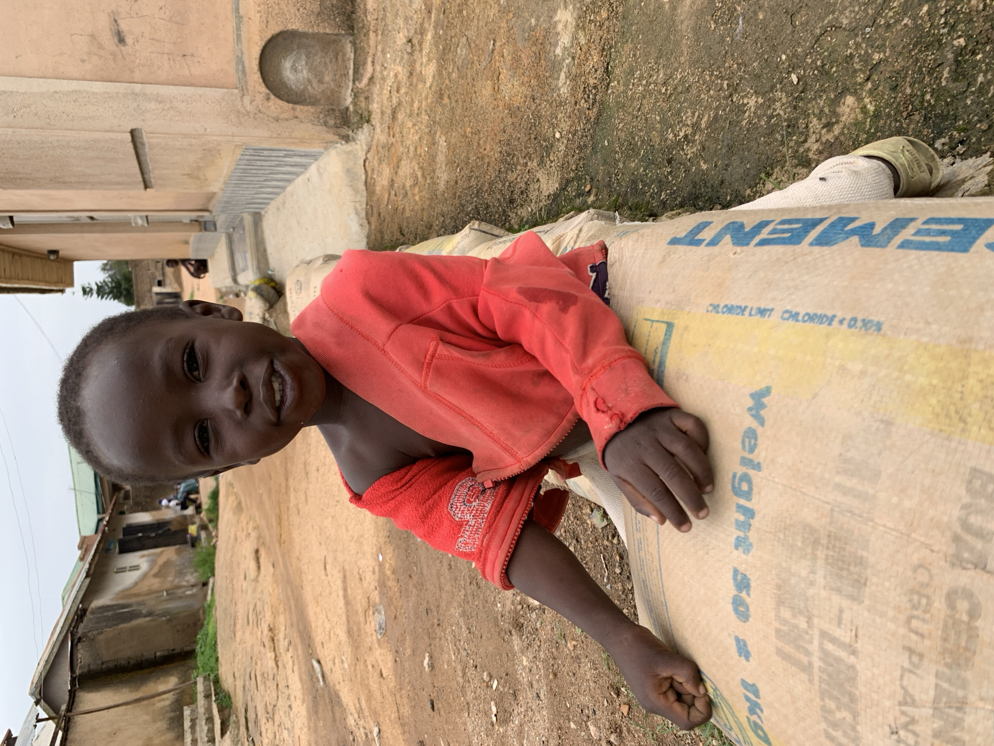 A young Black child sits on a bag of cement in front of a weathered building in Jos, Nigeria. The child is wearing a red zip-up jacket, and their left arm rests on the bag. The bag of cement is light brown with blue lettering that reads "CEMENT." The background shows a dusty street with more buildings and some green vegetation. The sky is overcast.