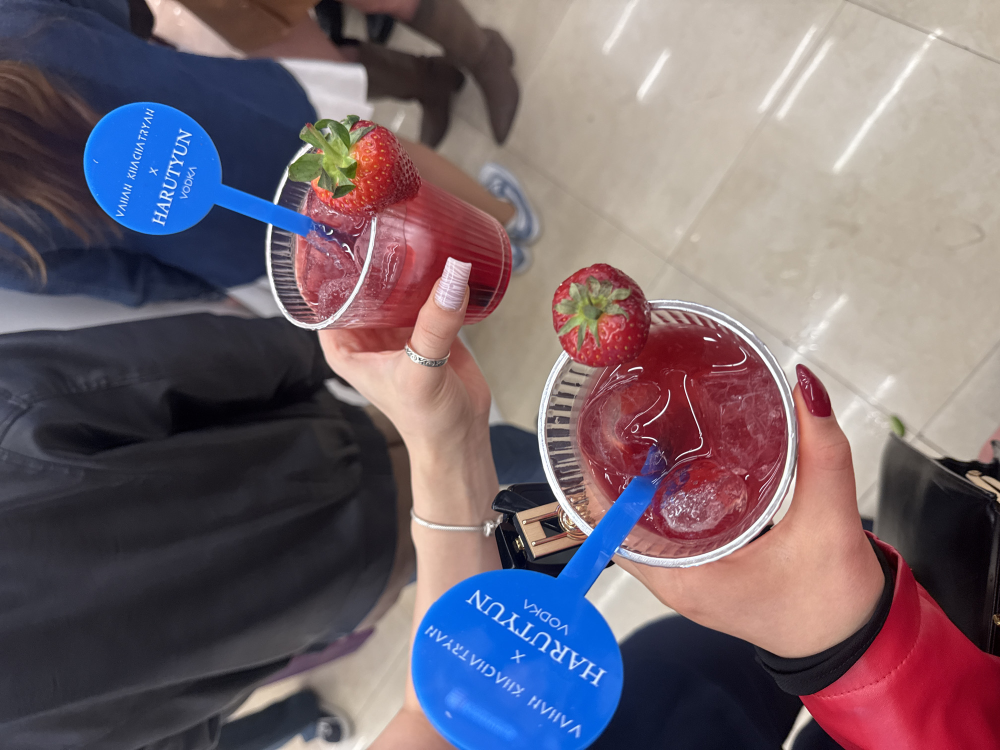 From an elevated perspective, the image captures two individuals each holding a red cocktail. Each cocktail is garnished with a strawberry and a blue plastic stirrer bearing white text, possibly a brand name or logo. The cocktail on the left is in a taller, cylindrical glass with a slight red tint, while the cocktail on the right is in a shorter, transparent plastic cup.
The person on the left has patterned nails, a silver ring, and a silver bracelet. The individual on the right has dark red painted nails. Clothing details include a black garment on the left and a red leather jacket with black cuffs on the right.
Background elements include other people, some wearing boots, and a glossy tiled floor. The setting might be indoors, possibly a bar, restaurant, or event. Given the details, it's impossible to definitively confirm the location as Yerevan, Armenia without additional information.