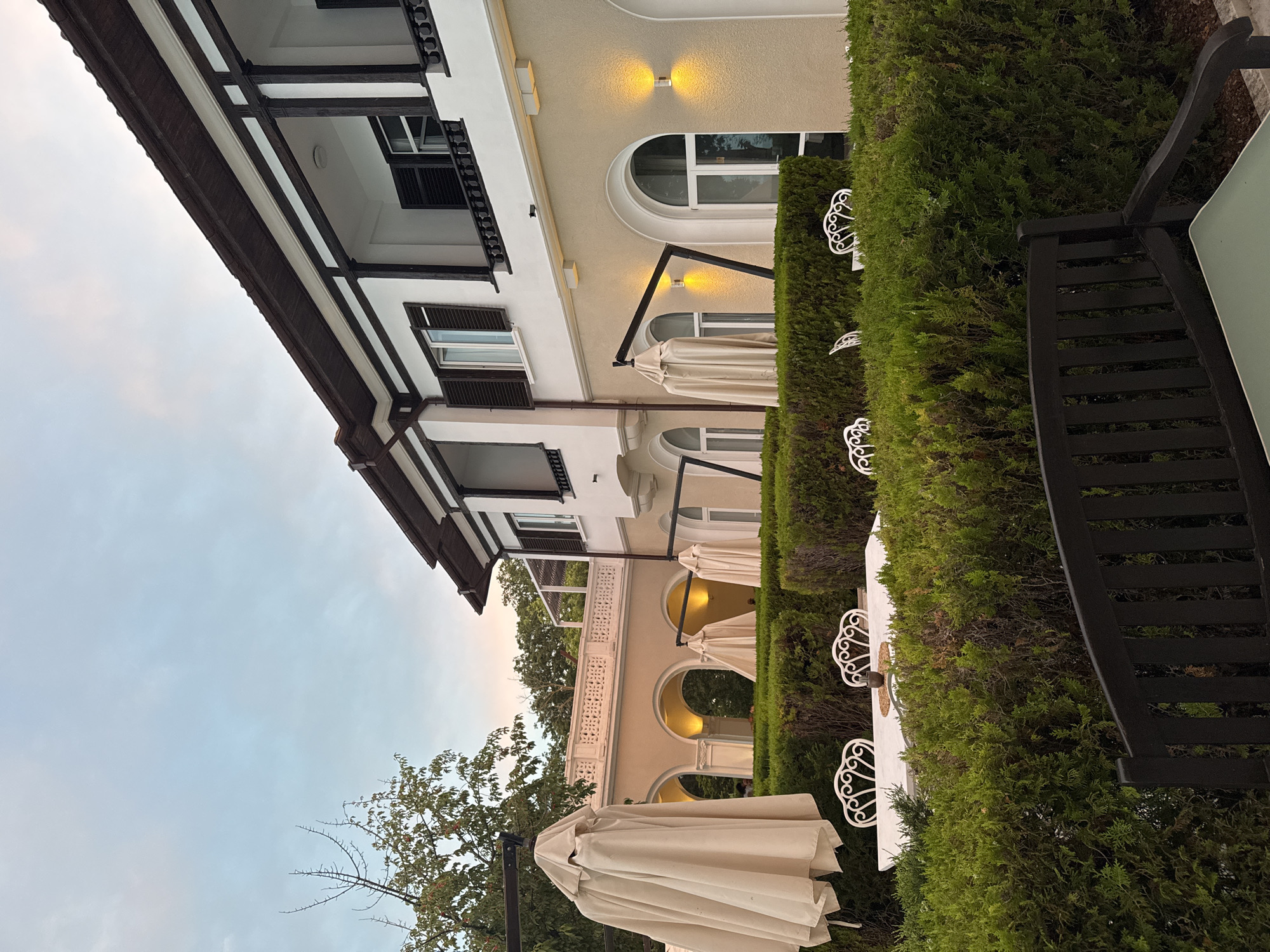 This image captures an outdoor seating area at the Grand Hotel Varna, situated in the resort complex of St. Constantine and Helena, near Varna, Bulgaria. The scene is set in the early evening, with a soft, twilight sky overhead.
In the foreground, a well-maintained hedge forms a green barrier, partially obscuring a white marble-topped table adorned with a small decorative item. Several ornate white chairs surround the table, hinting at a dining or relaxation space. To the left, a large, beige umbrella is folded and positioned on a metal stand, suggesting it is not currently in use. Beyond the hedge, the architecture of the Grand Hotel Varna is visible. A beige-colored building features a series of arched walkways with internal lighting, creating a warm glow. The upper levels of the building display a white facade with dark trim and balconies, featuring numerous windows, some with closed dark shutters. The building is set against a backdrop of lush green trees, typical of the coastal Bulgarian landscape. The overall impression is one of tranquility and refined outdoor leisure.