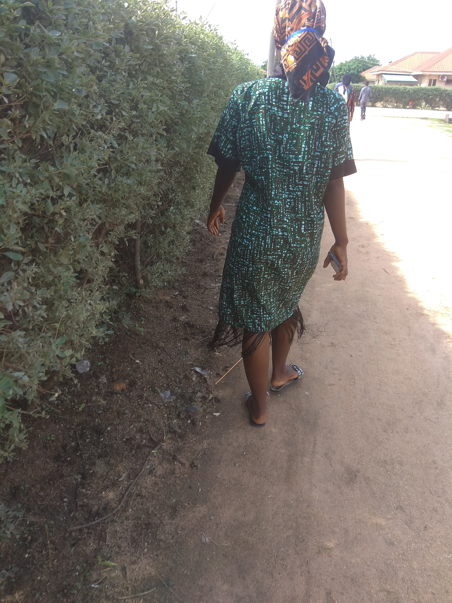 In Jalingo, Nigeria, a person walks away from the camera along a dirt path, bordered on the left by a dense green hedge and on the right by a sunlit area. The person wears a patterned green and blue dress with a fringed hem and patterned headscarf. Their right hand is extended slightly, holding a dark-colored mobile phone. Two other figures are visible in the background walking in the same direction. Buildings with terracotta roofs are discernible beyond the hedge. The ground is a mixture of dirt and sparse vegetation.