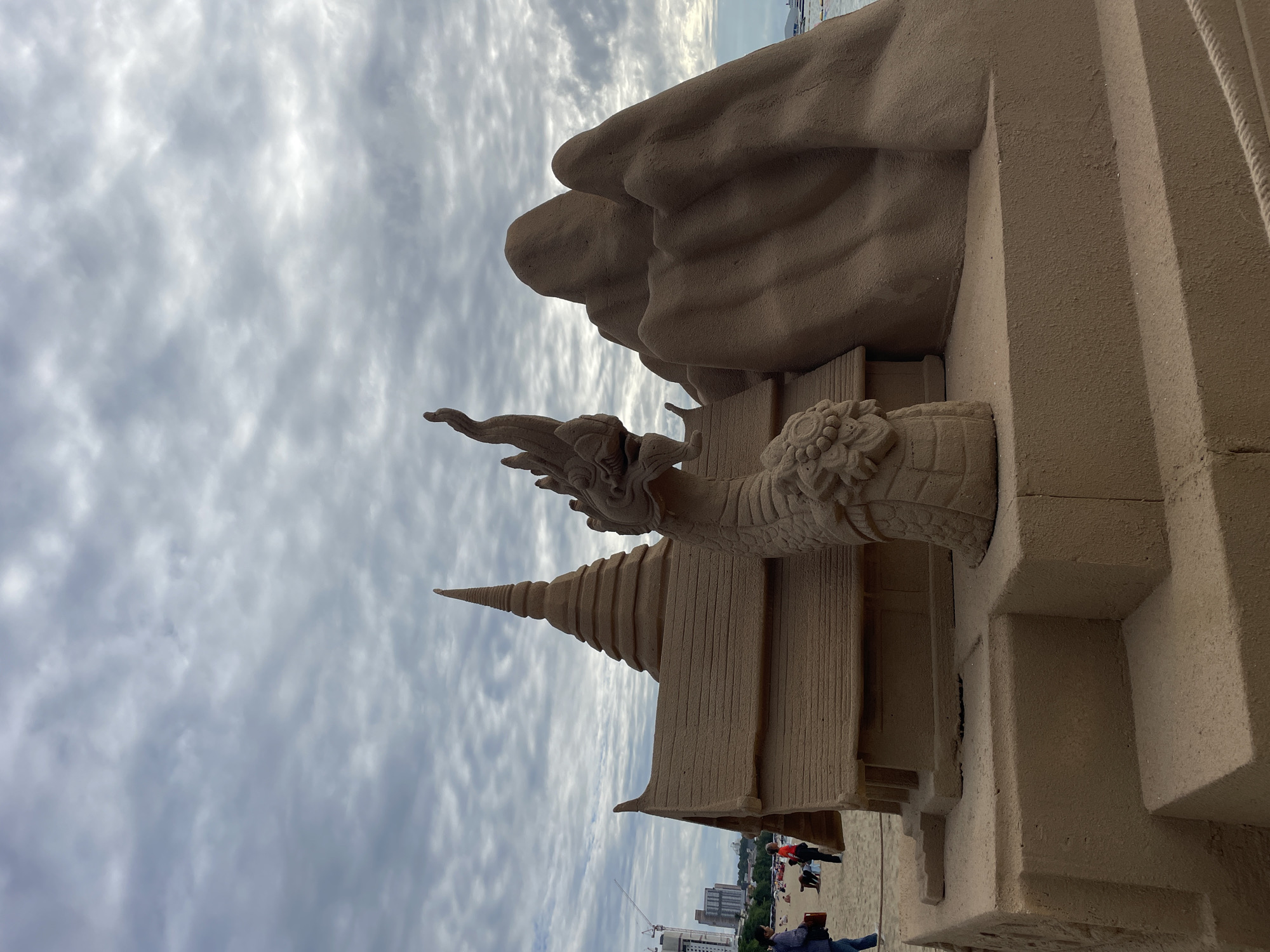 The image depicts a sand sculpture on a beach in Pattaya City, Thailand. The sculpture is intricate, featuring a temple-like structure with a tiered roof and a spire, accompanied by a Naga serpent statue in front of it. To the right of the temple, a sand sculpture of mountains is visible. The background shows a cloudy sky, the ocean, and distant buildings and cranes, indicating an urban coastal setting. Several people are scattered on the beach in the background, some walking and others sitting. The sculpture is positioned on a raised sand platform with steps. A rope railing is visible in the lower right corner of the image.