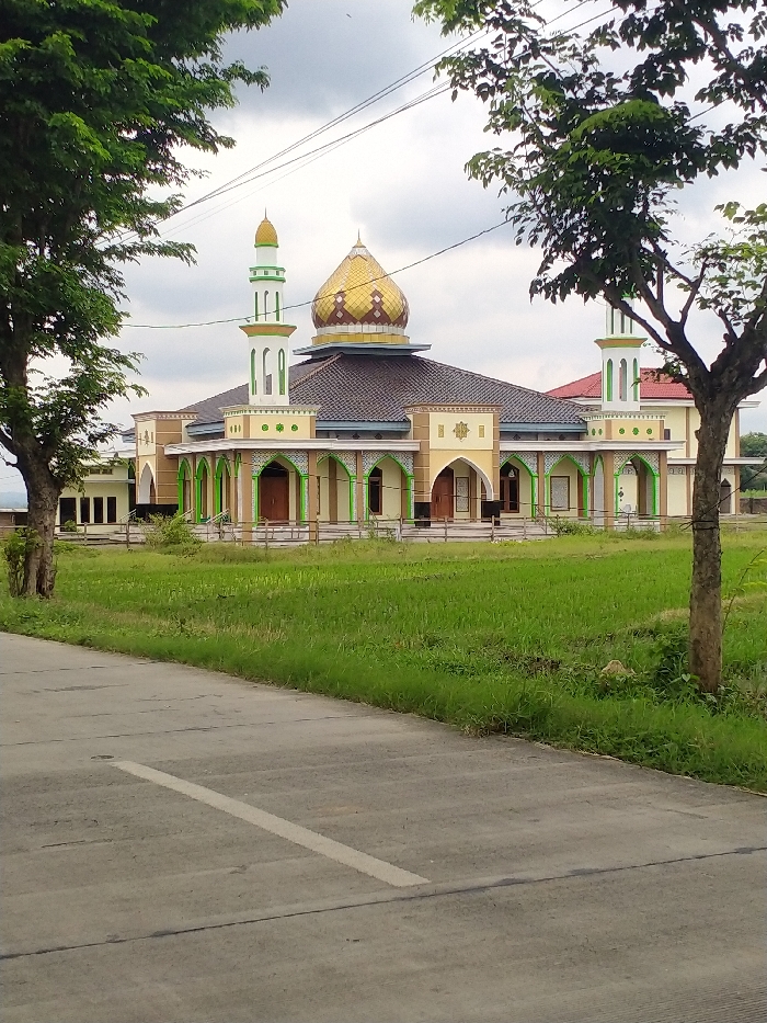 The image captures a modern mosque with a distinctive golden dome and two slender minarets, set against an overcast sky. The mosque features light yellow walls with green and brown decorative accents, including a series of arched facades along its front and sides. The roof is dark brown with a tiled appearance. The minarets are white and green, topped with small golden finials, complementing the larger central dome which has a patterned surface.
The mosque is framed by lush green trees on both the left and right sides of the foreground, their branches and leaves extending into the upper corners of the frame. In front of the mosque, there is an expanse of vibrant green grass or cultivated field. A paved road or path, light grey in color with a faint white marking, occupies the lower left portion of the image, curving away into the distance. Power lines are faintly visible stretching across the cloudy sky above the mosque.
The scene depicts a tranquil daytime setting, likely late morning or early afternoon, characterized by soft, diffused light due to the cloudy weather. No people or active events are visible. The architecture of the mosque and the surrounding natural landscape suggest a peaceful, possibly rural or semi-rural, environment in Kecamatan Kebakkramat, Indonesia.