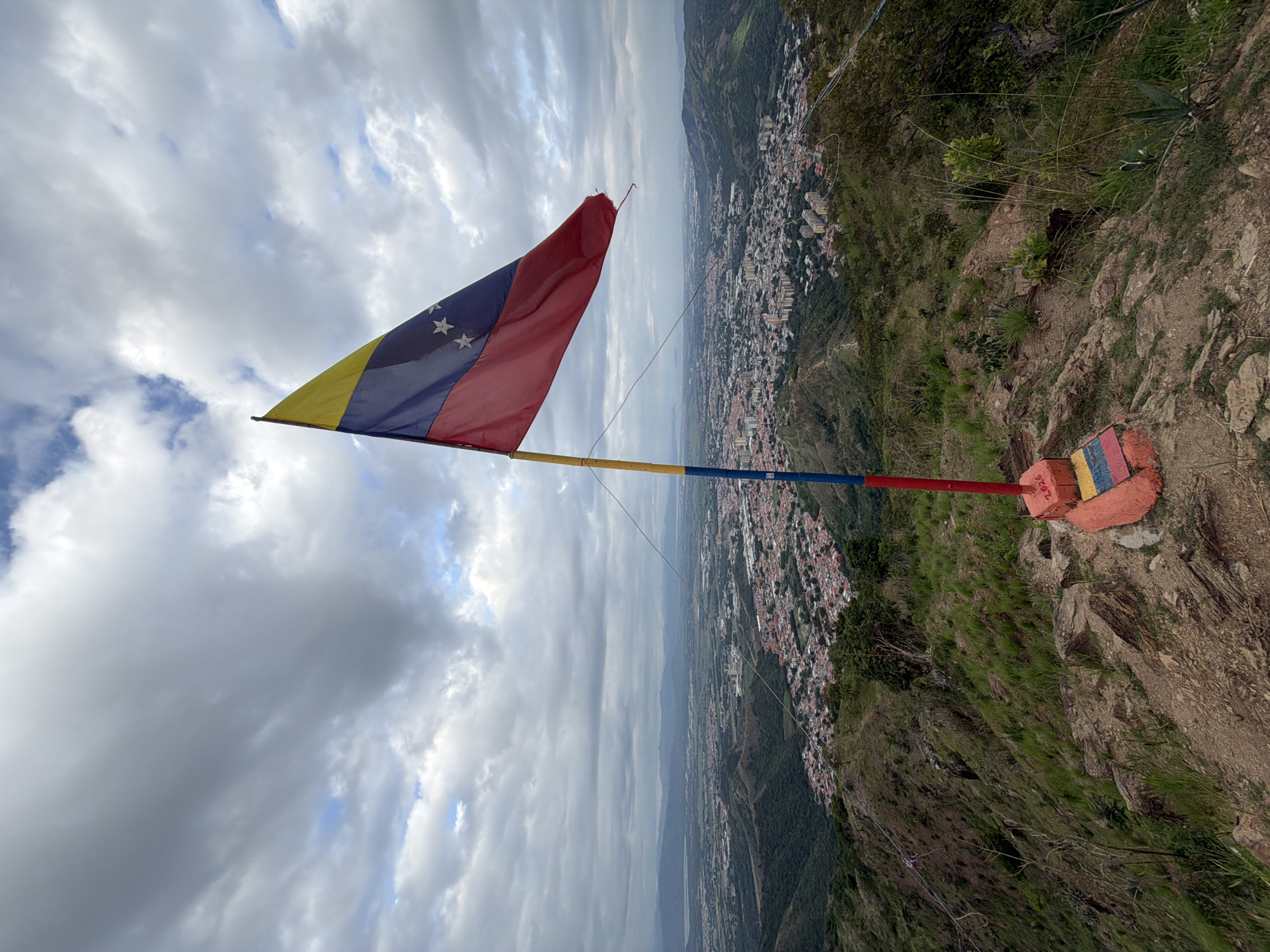 A Venezuelan flag flies from a pole on a hilltop overlooking a city under a cloudy sky. The flag, with its yellow, blue, and red stripes and white stars, is prominently displayed in the foreground. The pole is painted in sections of red, blue, and yellow, and stands on a small, colorful concrete base decorated with the colors of the Venezuelan flag and the year "2026". The hilltop is covered in dry grass and rocks, with some sparse vegetation. In the background, a sprawling city, identified as El Cambur, Venezuela, is visible with its red-tiled roofs and buildings extending into the valley. Mountains form a backdrop to the cityscape, and the sky is filled with dramatic, grey clouds. The overall scene suggests a panoramic view from a high vantage point.