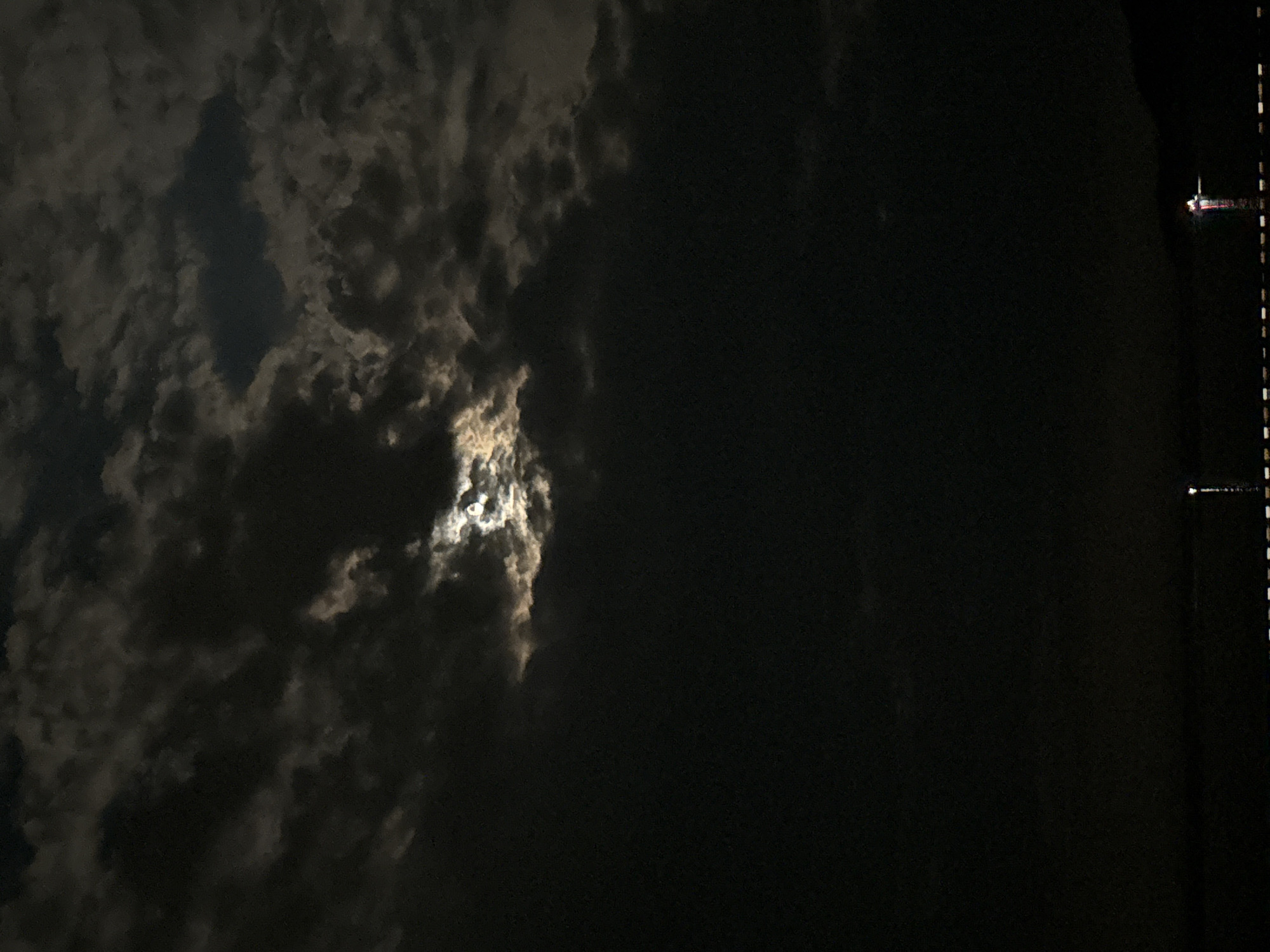 The image depicts a dark, cloudy night sky with a partially visible moon. The moon's light is diffused by thick clouds, creating a soft, ethereal glow that illuminates portions of the sky. Some clouds are dark and ominous, while others are tinged with moonlight, giving them a textured, almost sculpted appearance.
In the lower right portion of the image, a faint indication of a man-made structure or object is visible. It appears to be a vertical line with small, illuminated points along its length, possibly lights on a pier or a bridge. Further out in the dark expanse, a small, distant light source with a reddish hue is discernable, suggesting a boat or another vessel on the water.
The scene evokes a sense of quiet solitude and mystery, characteristic of a nighttime seascape. The dominant elements are the dramatic interplay of light and shadow within the clouds and the vast darkness of the night. The weather appears to be cloudy, with the moon struggling to break through. The presence of distant lights hints at human activity despite the overwhelming darkness and natural elements. No text is visible in the image.