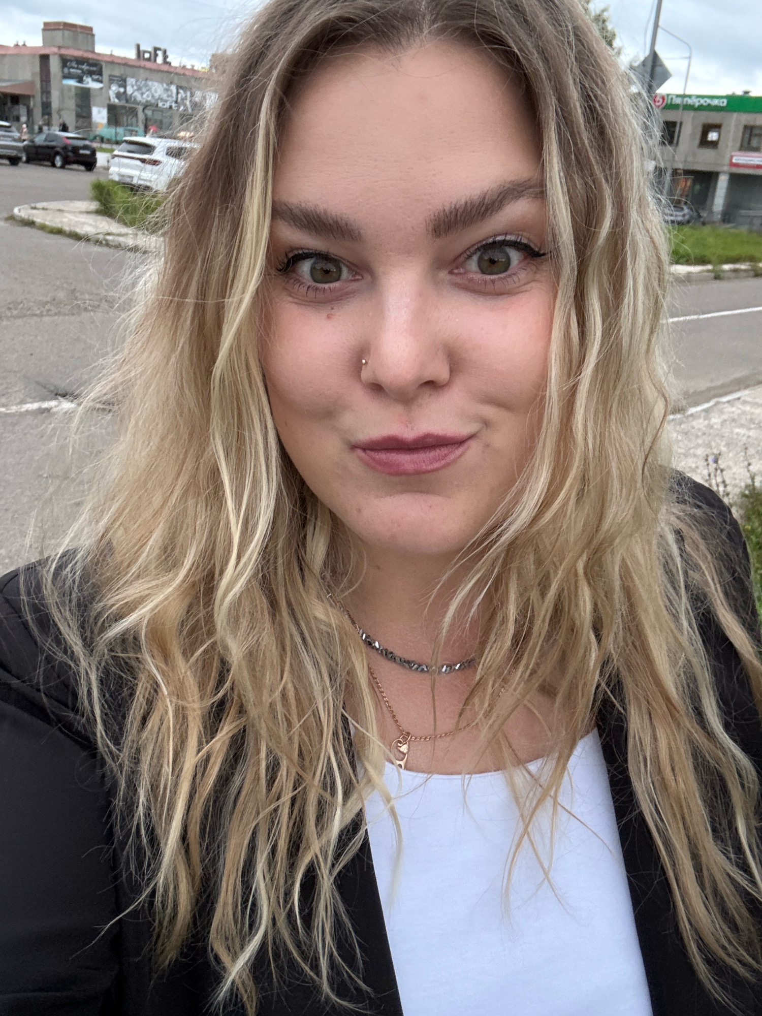 A woman with blonde hair is taking a selfie outdoors. She is wearing a black blazer over a white top and a necklace. Her hair is slightly messy and falls around her shoulders. The background shows a street scene with buildings, cars, and a cloudy sky. One of the buildings in the background has the word "LOFT" visible on it. The context of this location being in Марковский, Russia is implied.