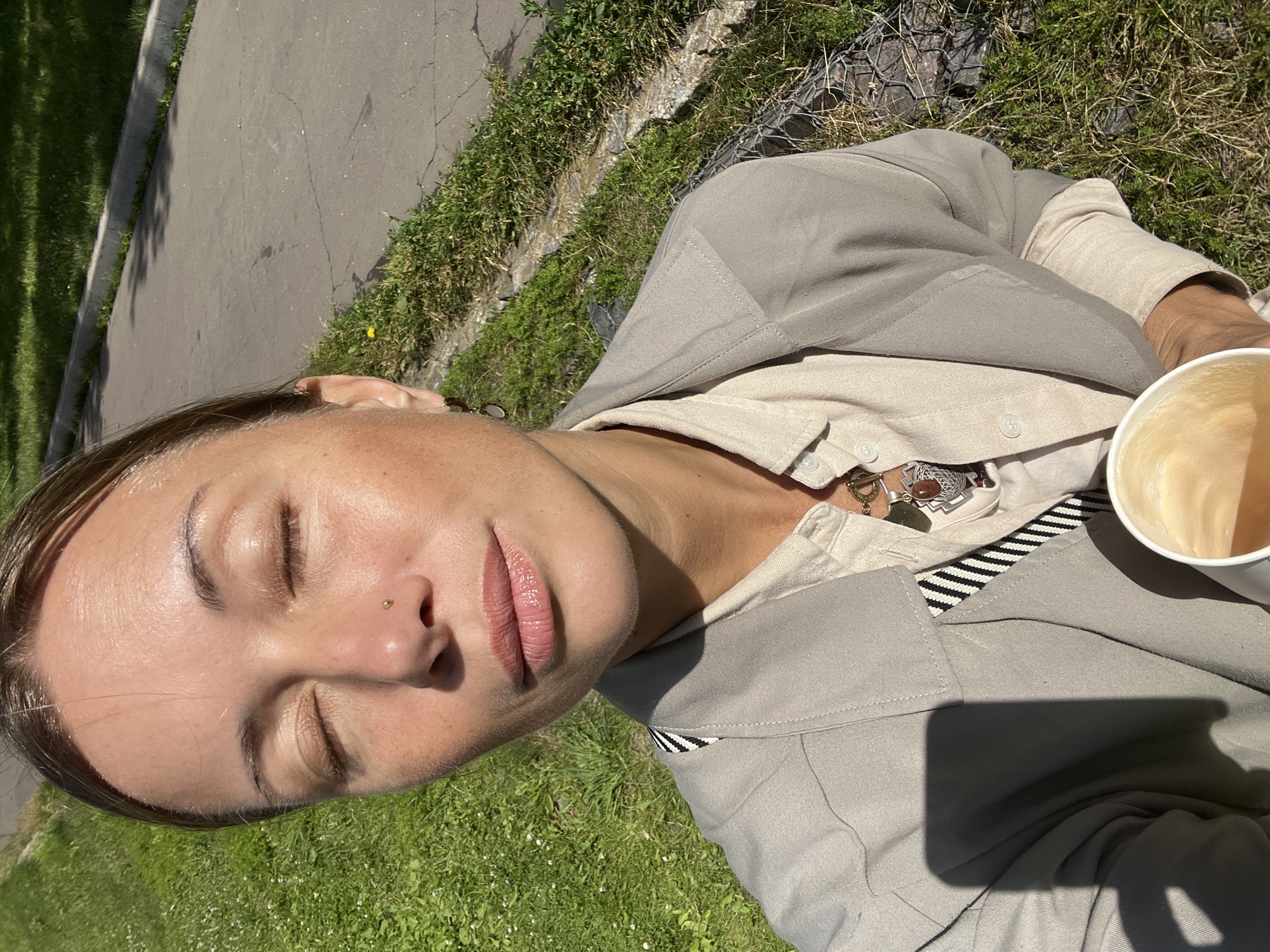 A woman with her eyes closed is in the foreground, her face angled slightly upwards. She is wearing a light-colored shirt and a grey jacket. A small stud is visible on her nose. Her lips are slightly parted and appear to be glossed. To her right, a white paper cup filled with a light brown liquid, likely coffee, is visible. The woman is outdoors, seated on a grassy area next to a paved surface that appears to be a sidewalk or path. The grass is green with some scattered white flowers. In the background, a wire mesh fence is visible, and beyond that, more grass and a hint of a building or wall. Sunlight is casting shadows, suggesting it is a sunny day. The image's orientation suggests the photo was taken in portrait mode. The presence of grass and the sunny weather imply a daytime setting, likely spring or summer.