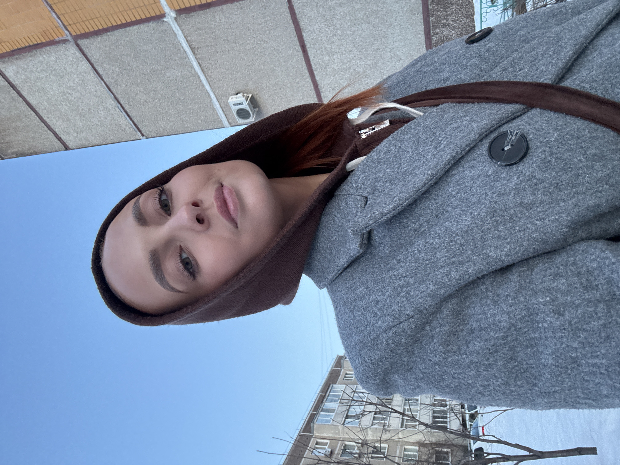 A female subject is captured in a slightly low-angle, head-and-shoulders portrait outdoors on a clear winter day in the city of Chaikovsky, Russia. She has reddish-brown hair, light eyes, and is looking directly at the camera with a neutral expression. She is wearing a dark brown hooded sweatshirt, with the hood pulled up, and a light gray textured double-breasted coat over it, fastened with large dark buttons. A white drawstring is visible from the sweatshirt.
The background features typical urban residential architecture. Immediately to the subject's right is a multi-story concrete building with horizontal structural lines and sections of orange-brown panels or cladding, typical of a modernized or re-clad Soviet-era building. An external air conditioning unit is visible on its facade. To the subject's left, in the mid-ground, is another multi-story light-colored apartment building with numerous windows and balconies. The ground is covered in snow, and several bare deciduous trees are visible in the mid-ground, indicating a winter setting. Partially visible behind the trees are a few parked vehicles. The sky above is clear and blue.