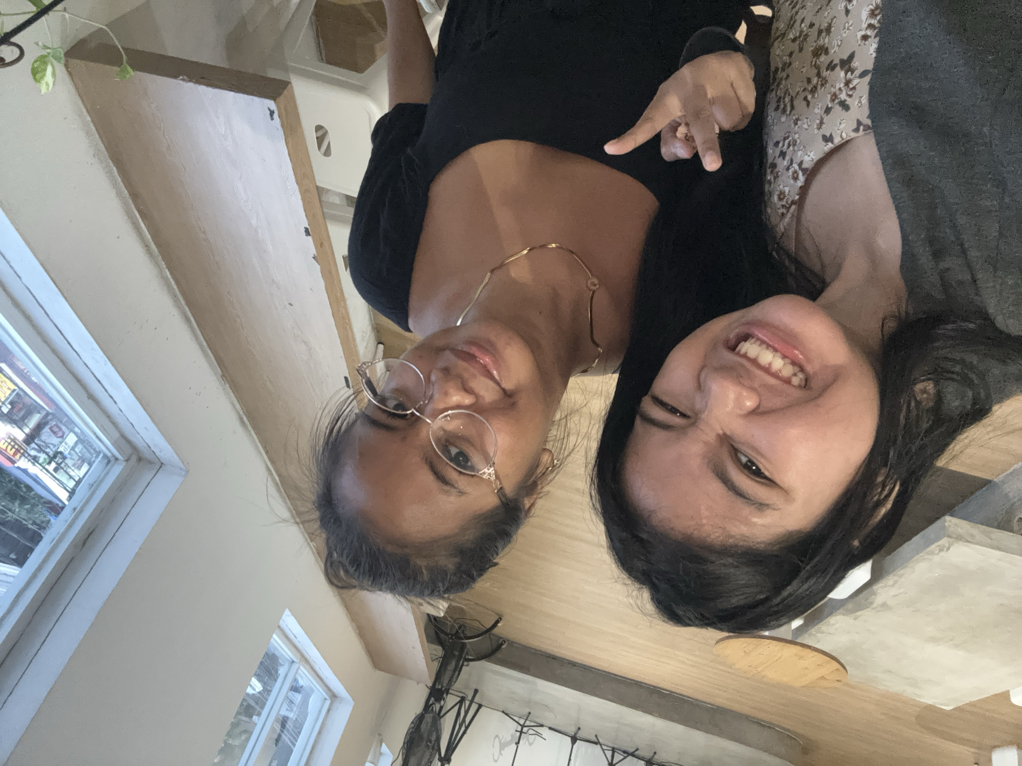 Two young women are posing for a selfie indoors. One woman is wearing glasses, a black top, and a gold necklace. She is pointing toward the other woman, who is wearing a floral top and a gray sweater. The background shows a window with daylight, interior walls, and a table with chairs. The location is in North Jakarta, Indonesia, as evidenced by street scenes visible through the window.