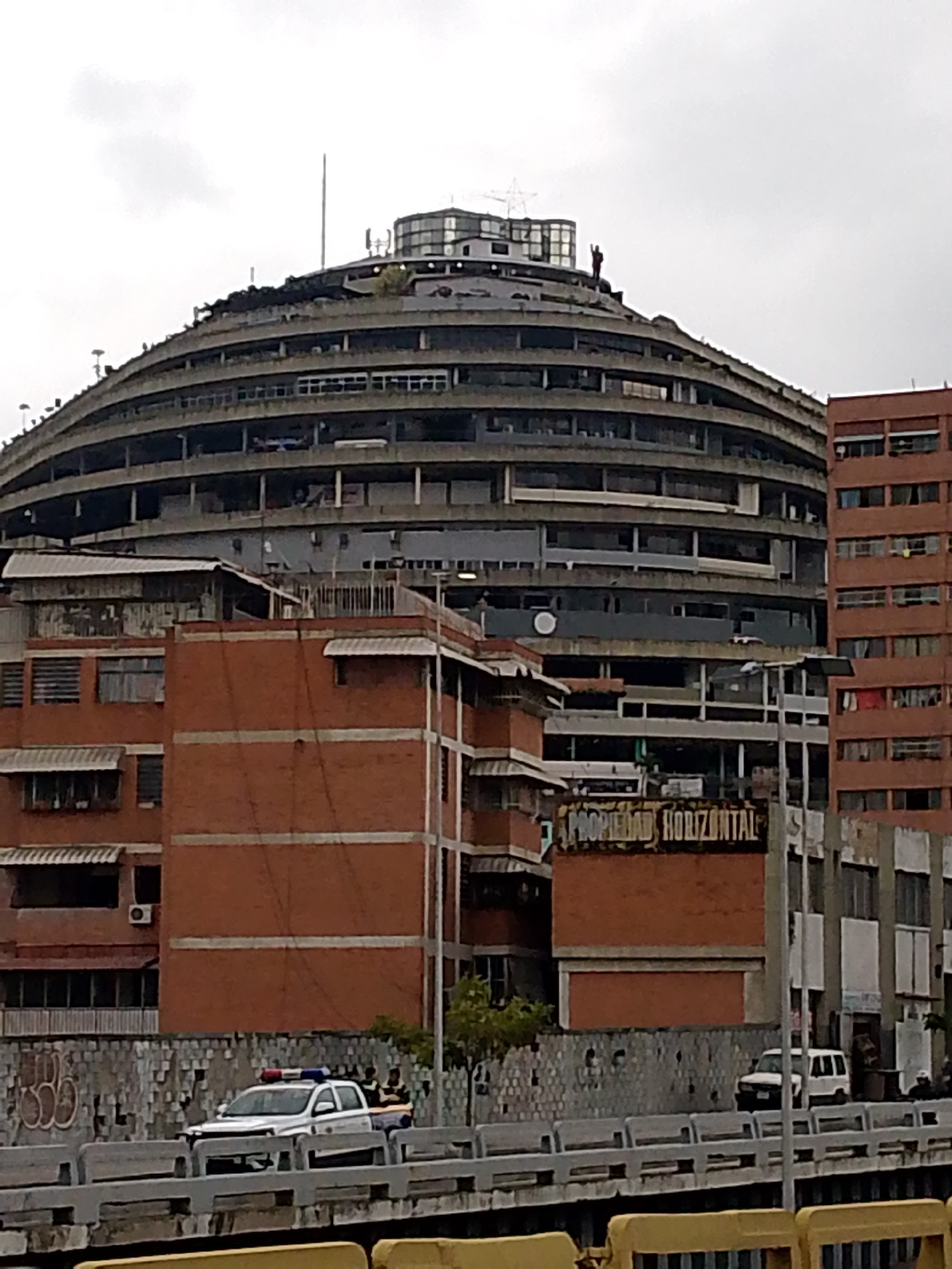 A wide shot captures an overcast day in the city of Caracas, Venezuela, focusing on a large, distinctive circular building with multiple levels. The building's architecture features a repeating pattern of balconies or open-air floors, with concrete structures dominating its design. Smaller, reddish-brown brick buildings are situated in front of the circular structure, some with awnings over windows. A sign is visible on one of the brick buildings reading "PROPIEDAD HORIZONTAL". In the foreground, a white police vehicle with red and blue lights is parked on a bridge or elevated roadway. Other vehicles, including a white SUV, are visible further down the road behind the police car. A low, textured wall with graffiti separates the road from the buildings. The sky is uniformly gray and cloudy, indicating a dull or potentially rainy day.