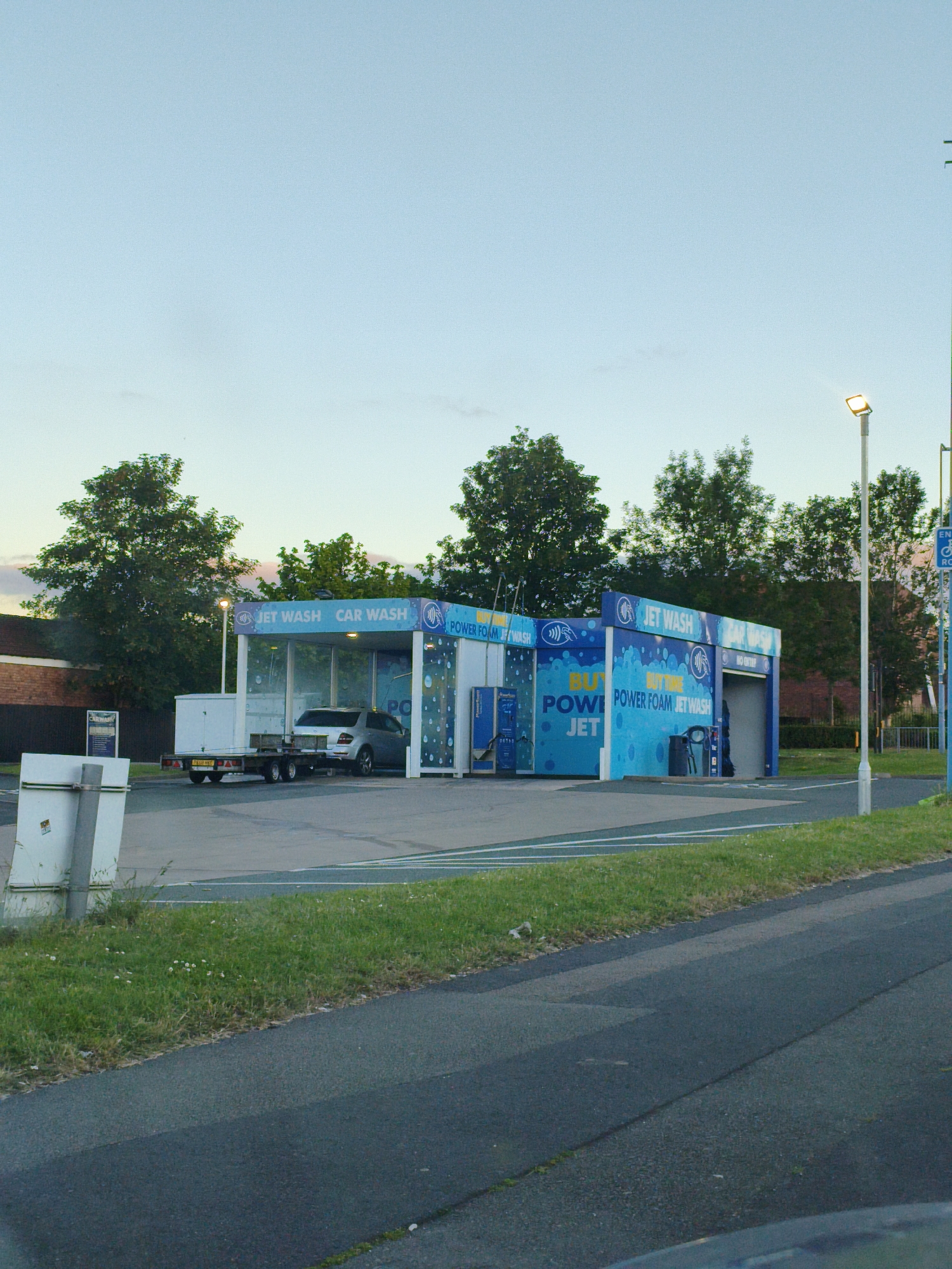 In Sandwell, United Kingdom, at dusk, a car wash facility is visible. The facility is a modern, open-air structure with blue and white branding. The words "JET WASH" and "CAR WASH" are prominently displayed on the building. A silver sedan is positioned within the wash bay, and a trailer is attached to the rear of a white box truck parked nearby. The area surrounding the car wash is paved, with a grassy verge and a road in the foreground. A tall lamppost with a lit bulb stands to the right of the car wash, illuminating the scene. Trees form a backdrop to the car wash, and a brick building is visible in the distance. The sky is a pale blue, indicative of twilight.