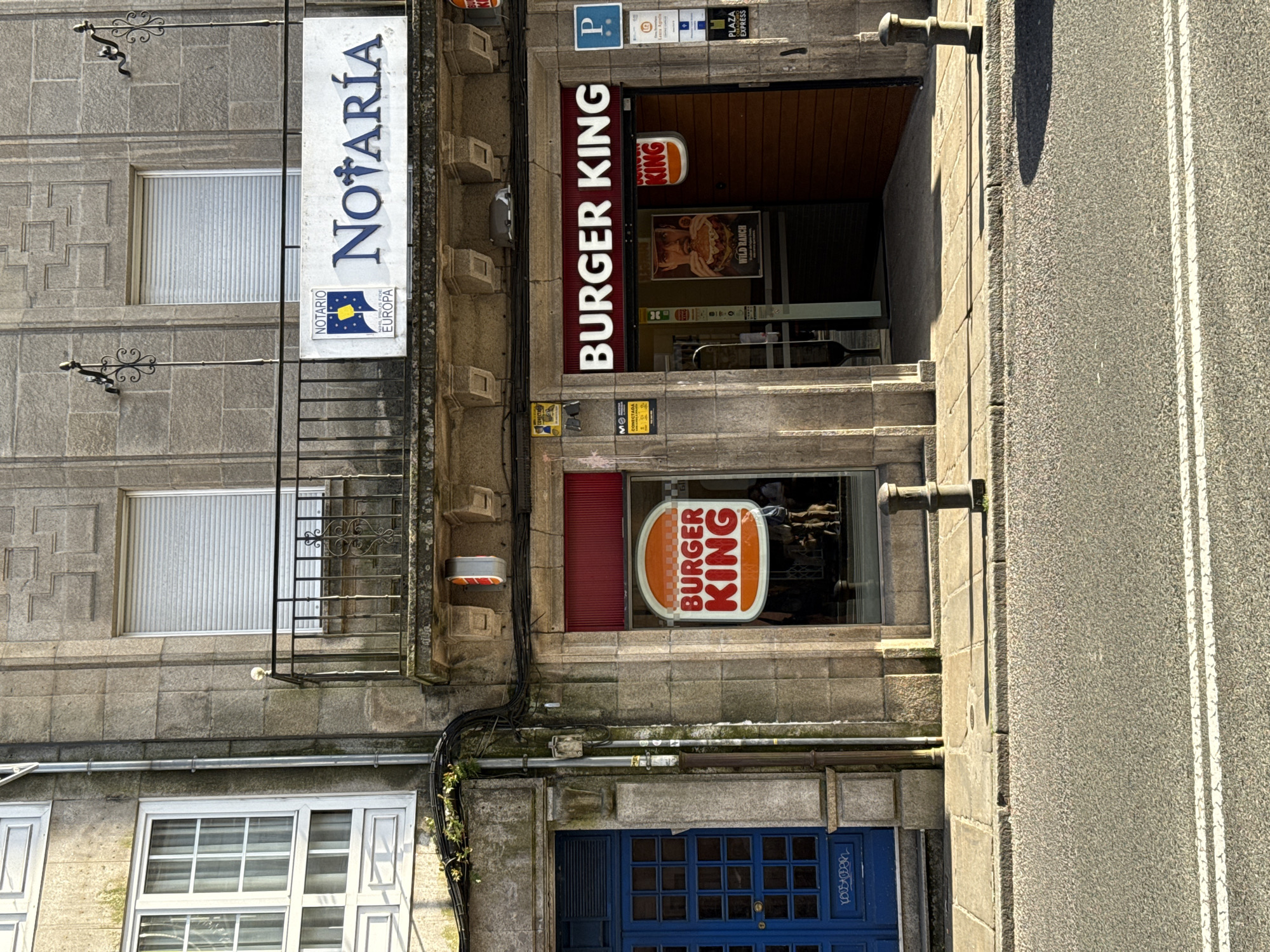 The image depicts the façade of a building in Santiago de Compostela, Spain, housing both a Burger King restaurant and a notary's office ("Notaría"). The building is constructed of gray stone.
On the upper floors, a sign for the "Notaría Europa" is vertically displayed. The building features windows with white frames and ornate wrought iron balconies.
On the ground floor, a Burger King restaurant is identifiable through its signage. A rectangular sign reading "BURGER KING" is visible above the entrance, along with the Burger King logo on the front window. The interior features fast food advertisements. A parking sign is also shown in the image. Below the Burger King, a blue door with small, square windows is visible.
To the right of the building, part of a street and sidewalk is visible. The street has painted white lines marking the lanes. The overall impression is of an older building adapted to commercial use, with fast-food and legal services coexisting in a historic urban setting.
