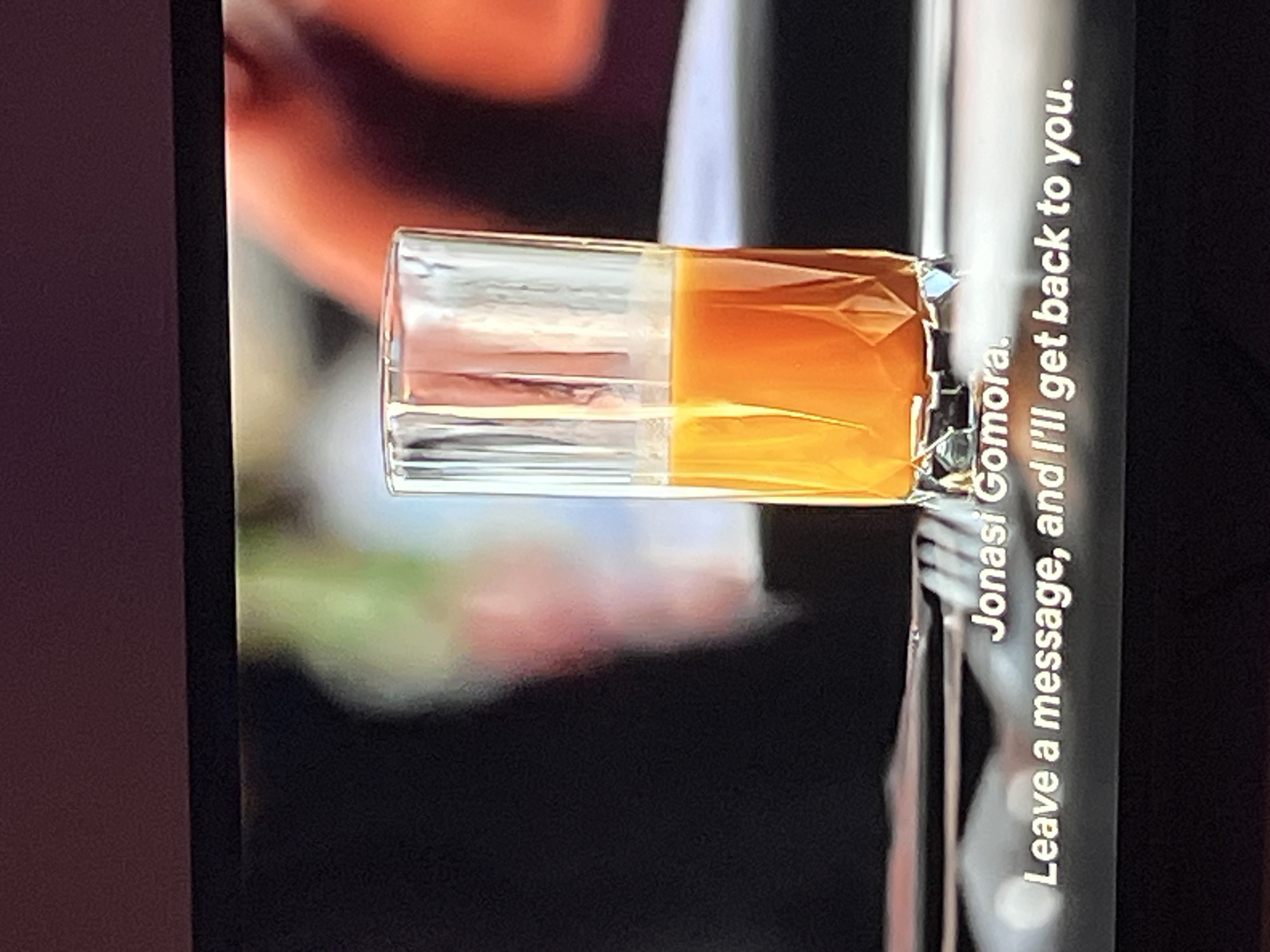 A close-up shot features a clear shot glass filled with an amber liquid, split halfway between clear and orange, suggesting a layered drink. The glass is placed on a reflective surface. To the right of the glass, white text runs vertically, reading "Jonasi Ggömora. Leave a message, and I'll get back to you." The background is blurred, with hints of orange and green, indicating an indoor setting or a bokeh effect. The image's lighting is soft, highlighting the textures of the glass and the liquid. The text suggests a communication prompt within the context of the visual.
