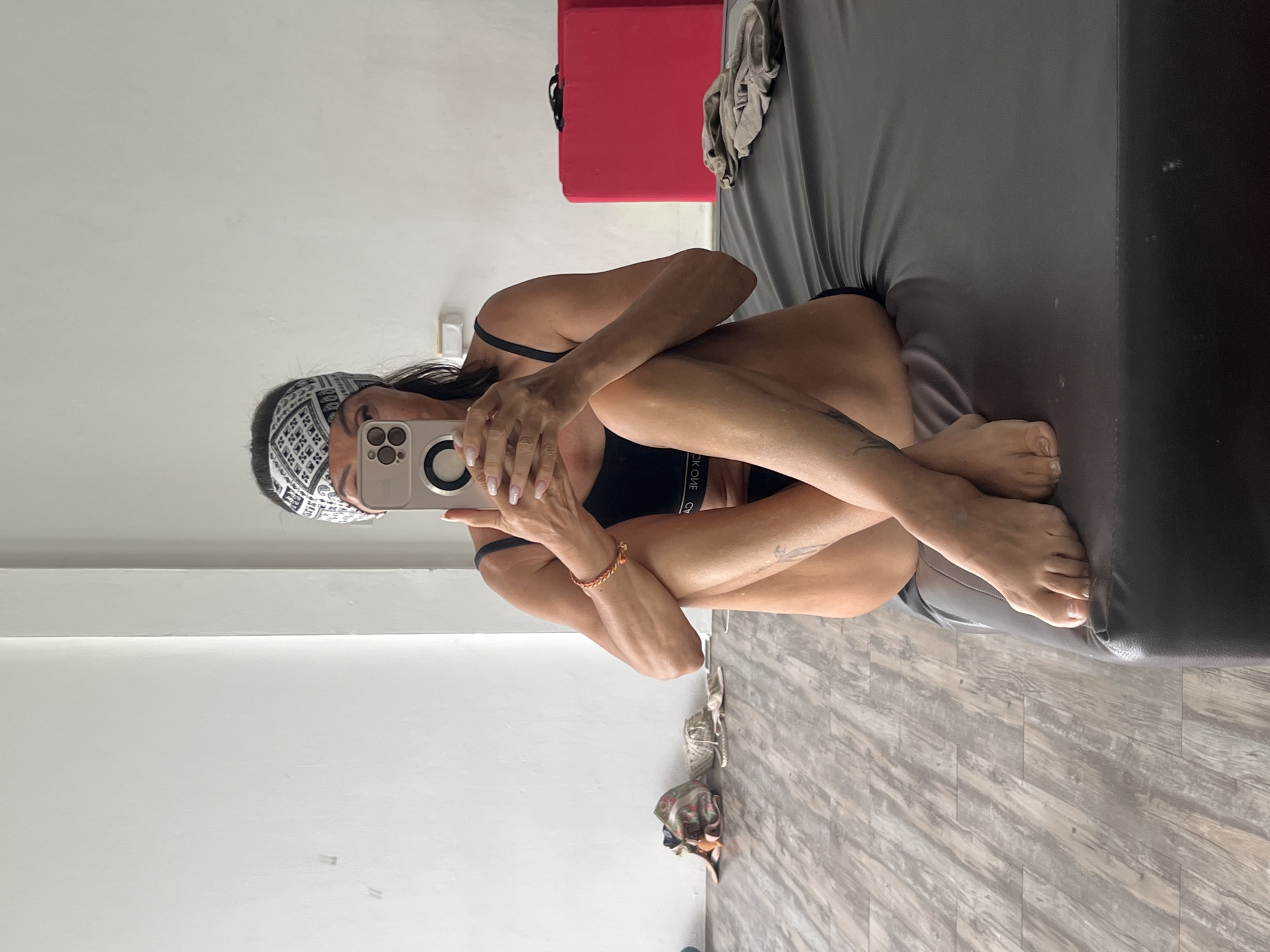 A woman sits on a bed, taking a selfie. She wears a black sports bra and a patterned headband. Her legs are crossed and drawn up to her chest. The setting is a room with gray wood-patterned flooring and white walls. A red object is visible in the background. Based on the location given, this is likely in บ้านบางรักษ์, Thailand.