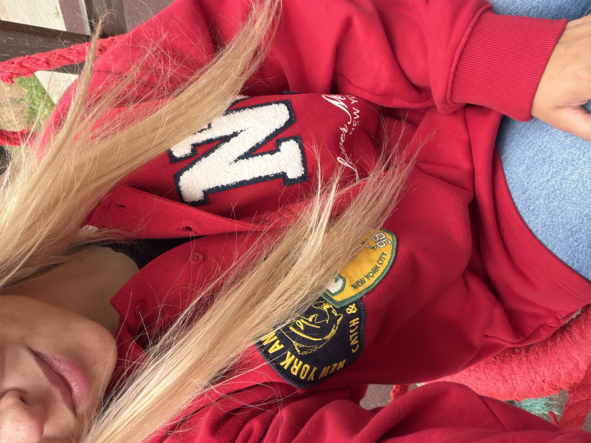 The image shows a close-up of a person wearing a red hooded sweatshirt with patches and text. A large white letter "N" with a dark blue outline is visible on the right side of the sweatshirt. A patch with yellow lettering that reads "NEW YORK AN" and a fish graphic is on the left. Below that, another patch with "CATCH &" and "NEW YORK CITY 98" is visible. The person has blonde hair that partially obscures the sweatshirt. The lower part of the image shows light blue jeans. The background is blurred but appears to be outdoors with some greenery and wooden structures. There is no indication of the city of Votkinsk, Russia in the image.