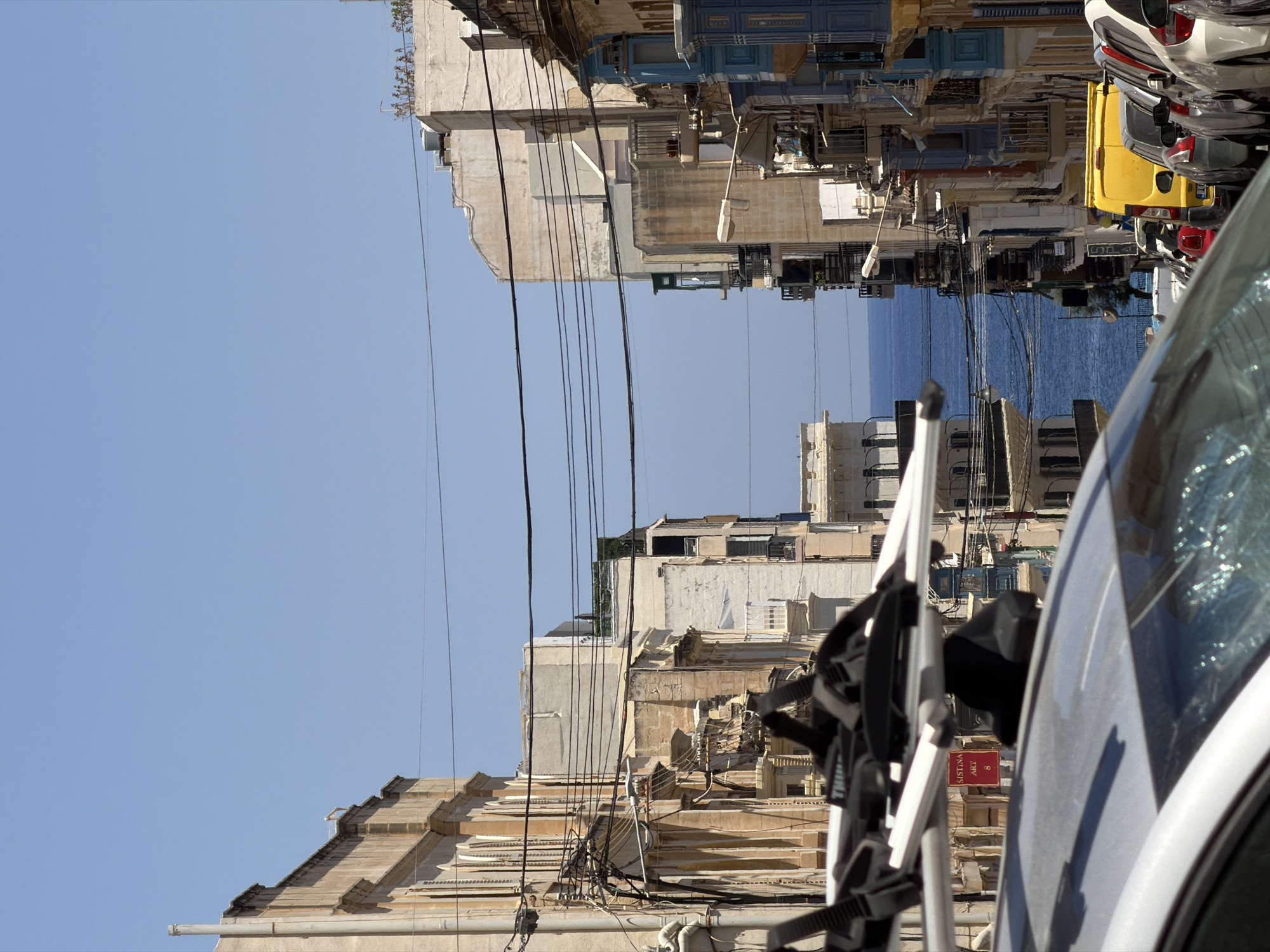 The image depicts a street scene in Sliema, Malta, looking towards the sea. In the foreground, the roof and windshield of a silver car are visible, with a bike rack mounted on top. Behind the car, a narrow street lined with multi-story buildings stretches towards the horizon. The buildings on both sides of the street feature traditional Maltese architecture, with balconies and windows. Power lines crisscross the sky above the street. The street itself is filled with parked cars, including a bright yellow van. In the distance, the blue of the Mediterranean Sea is visible between the buildings. The sky is clear and blue. A red sign with white text reading "SISTINA ART 8" is attached to the bike rack.
