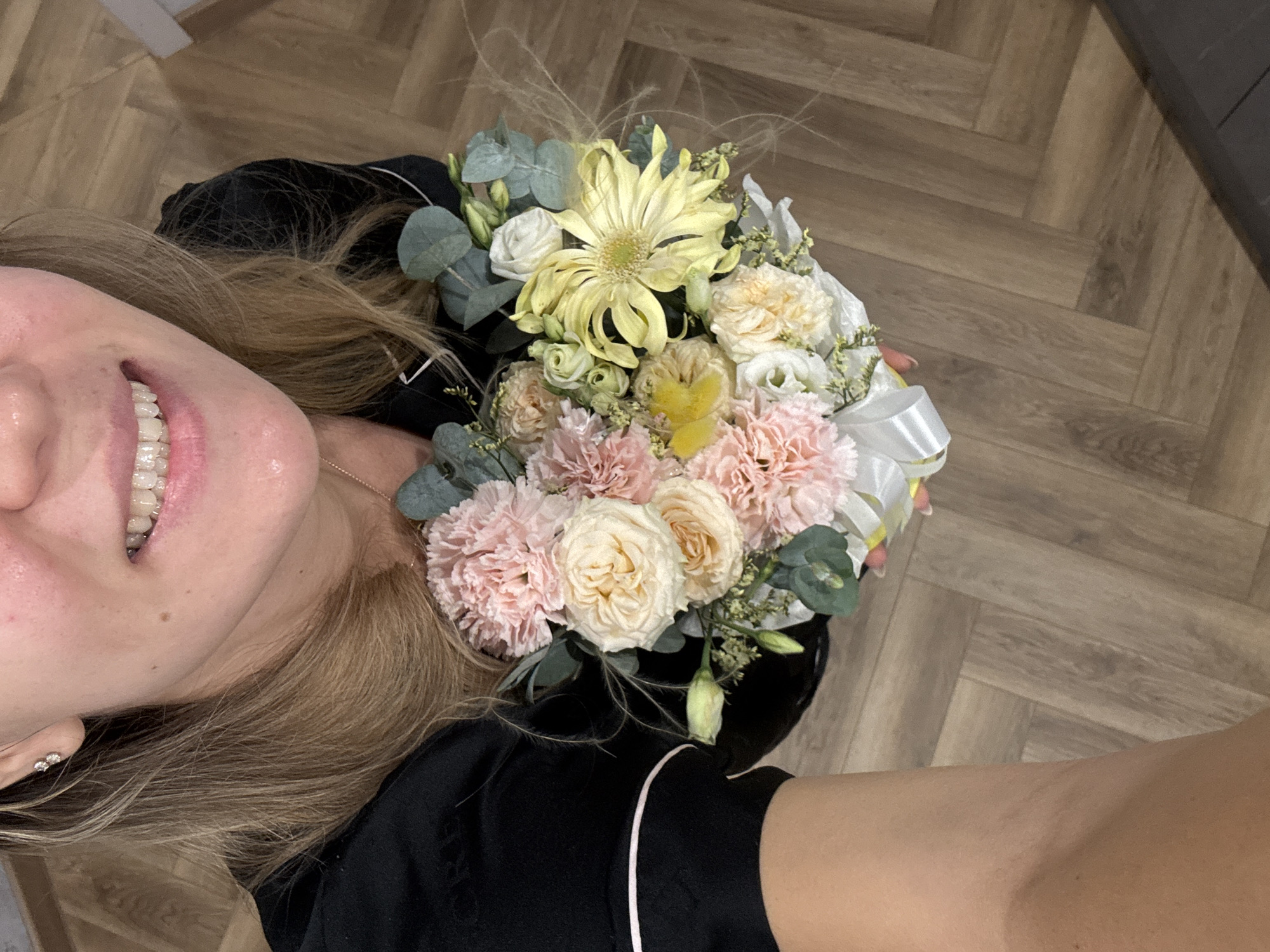 A woman is holding a bouquet of flowers in an indoor setting with a herringbone pattern wood floor. The bouquet features a yellow gerbera daisy as the focal point, surrounded by pink and cream-colored carnations and roses, along with eucalyptus leaves and other filler greenery. The flowers are tied with a white ribbon. The woman is wearing a black top with white piping. Her hair is blonde, and she is smiling, revealing her teeth, which have clear aligners. An earring is visible on her left ear. The location is an indoor space, but the specific city and region are unknown.
