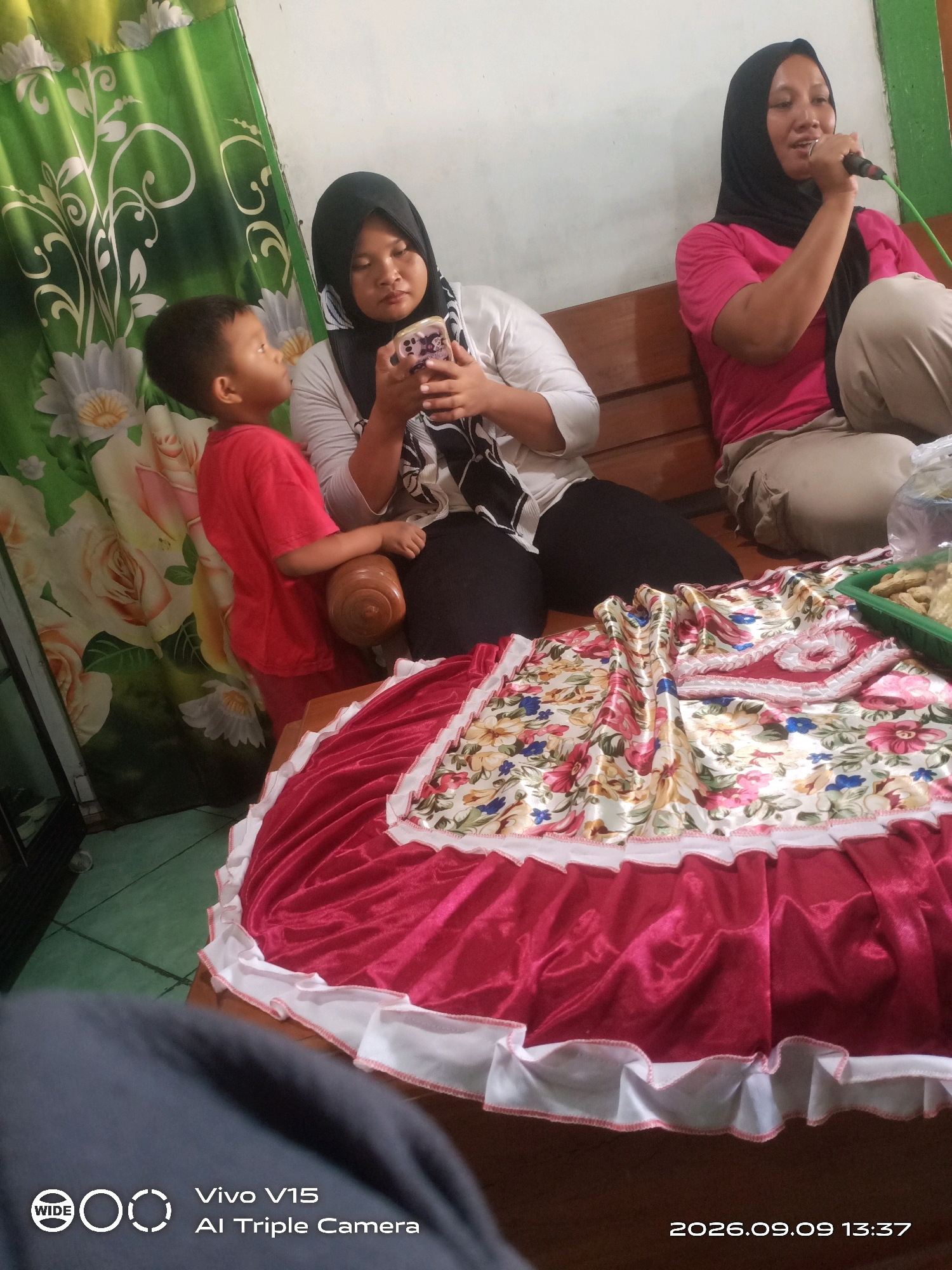 The image depicts an indoor scene likely within a residence in Plosorejo, Indonesia. Three individuals are visible: a young boy in a red t-shirt and pants standing on the left, looking upwards; a woman in a black hijab and white top sitting in the middle, holding a smartphone; and another woman on the right, wearing a black hijab and a pink t-shirt, holding a microphone to her mouth. They are seated on a wooden bench. In the foreground, a large, ruffled skirt with a floral print and a red satin underskirt is spread out. A green plastic container filled with food items, possibly snacks, is placed to the right of the skirt. The background features a green curtain with a floral pattern. The lighting suggests an indoor environment, possibly during the daytime. The bottom right corner of the image displays a timestamp: "2026.09.09 13:37" and a Vivo V15 AI Triple Camera watermark.