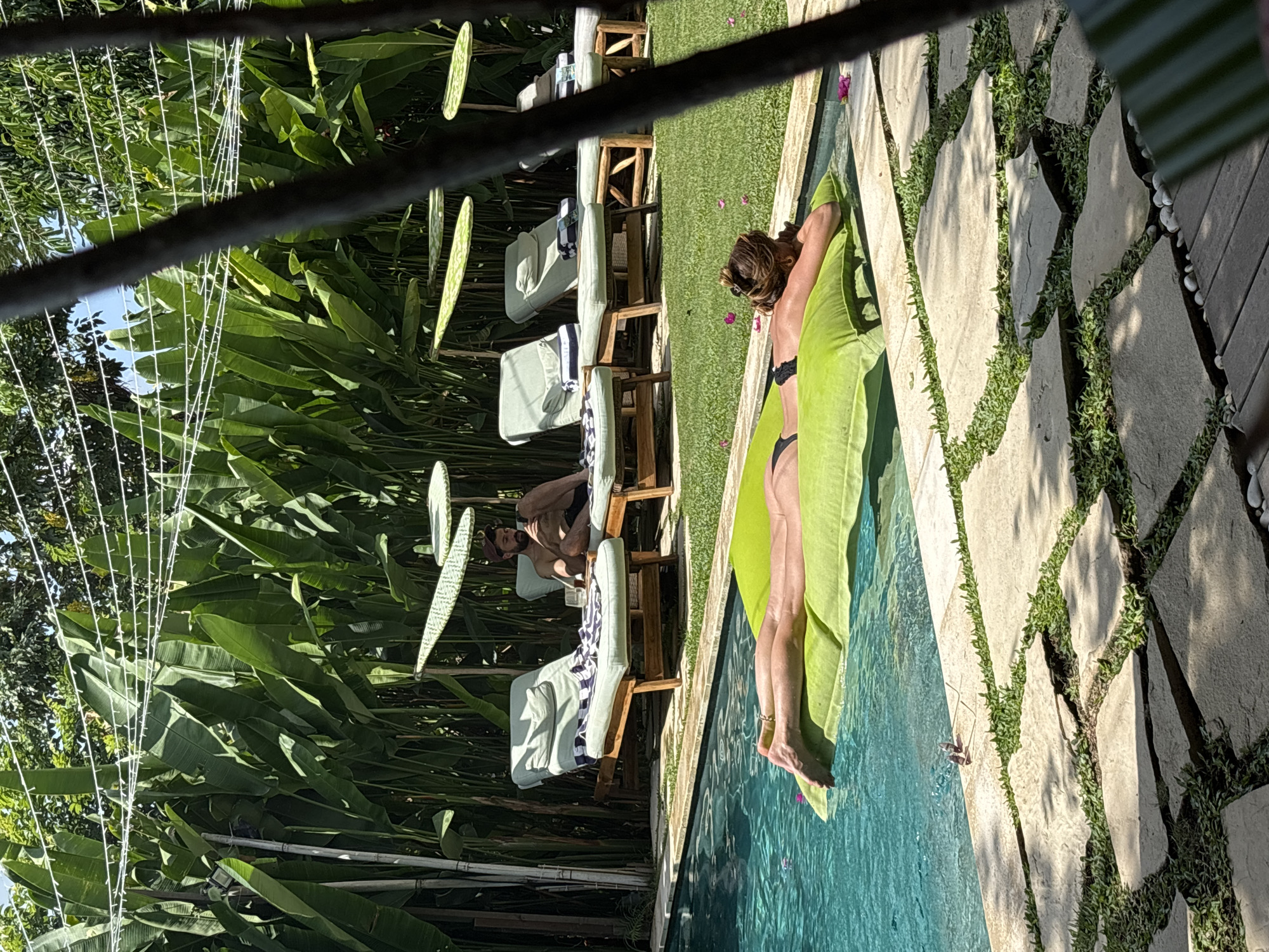 A daytime exterior shot shows a pool area in Kuta Selatan, Indonesia. A woman in a black bikini lies on a green towel at the edge of a pool, while a man sits on a lounge chair in the background. The area is lush with green foliage and a stone path borders the pool. Lounge chairs line the path. A string of lights hangs overhead.
