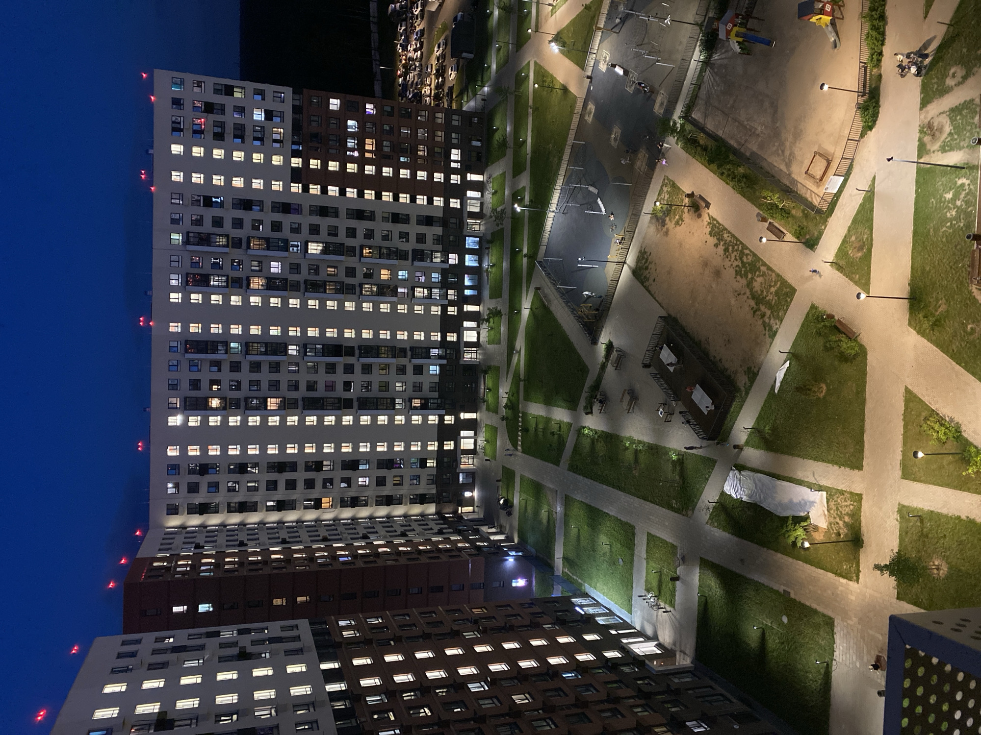 The image depicts a nighttime view of a residential complex in the city of Mirny, Russia. The complex consists of several tall, multi-story buildings with numerous windows lit from within. Red warning lights are visible on the rooftops of some of the buildings, suggesting an aviation hazard or navigational aid. The courtyard area between the buildings features paved walkways, grassy lawns, streetlights, and benches. There are also recreational areas, including a playground with slides and climbing structures, and a sandpit. Several cars are parked in a lot to the right of the buildings. A few individuals are visible in the courtyard and on the playground, with some appearing to be walking or playing. The overall scene conveys a sense of urban residential life at night, with the artificial lighting of the buildings and grounds contrasting with the dark blue twilight sky.