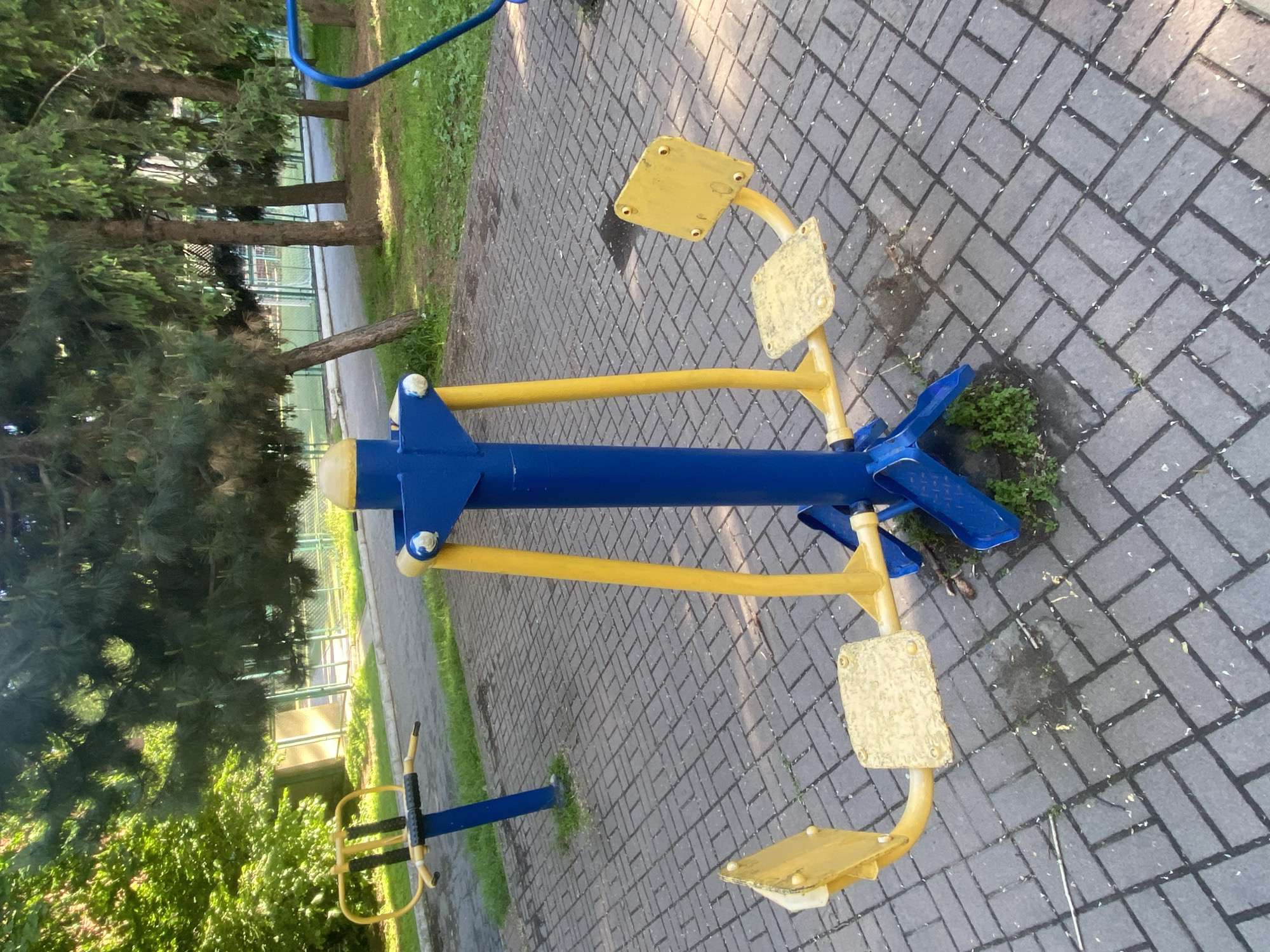 The image depicts outdoor exercise equipment in what appears to be a park setting, likely in Poltava, Ukraine. The equipment is predominantly blue and yellow. The exercise machine features two yellow foot rests on rotating yellow levers connected to a central blue mechanism. The ground surface is paved with gray bricks. In the background, trees and greenery are visible, suggesting a park or recreational area. A second, smaller exercise machine can be seen in the lower left, but it is more obscured. The lighting indicates it is daytime.