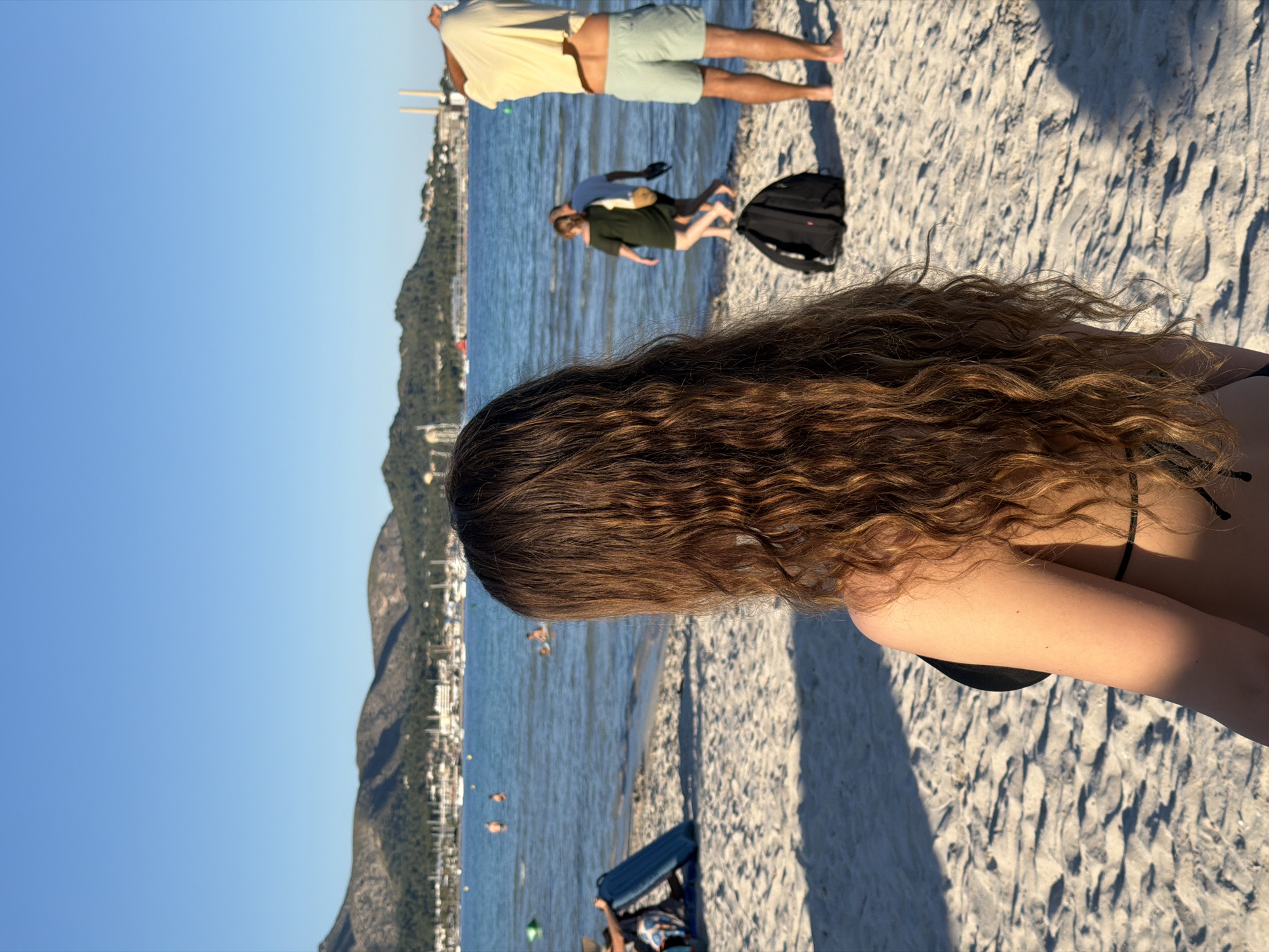 The image shows a beach in Alcúdia, Spain. A woman with long, wavy brown hair is in the foreground, facing the water. She is wearing a black bikini top. In the background, several people are enjoying the beach and the water. There are mountains and buildings visible on the horizon. The sky is clear and blue. The sand is light-colored and textured.