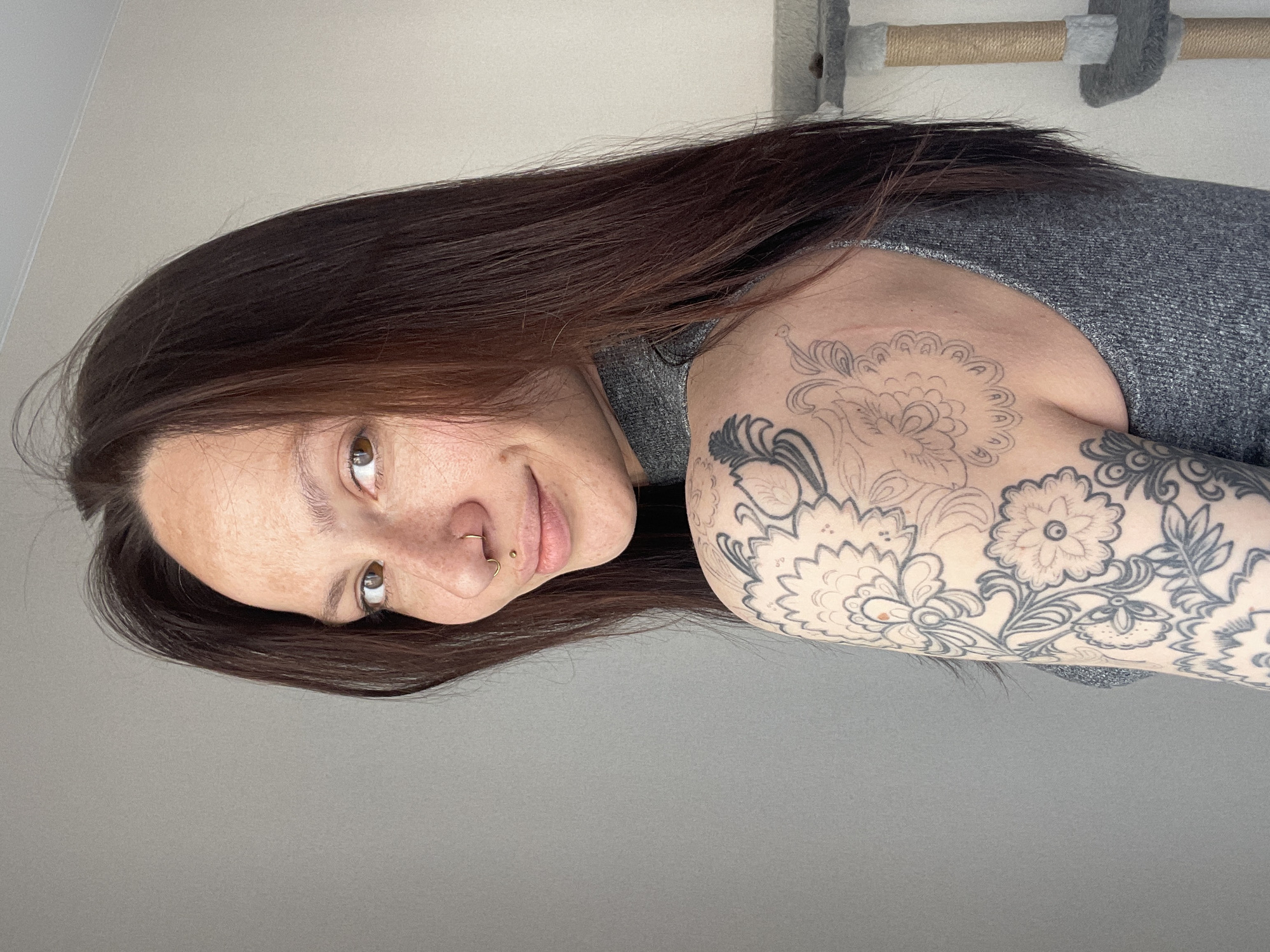 A medium shot shows a woman with dark brown hair. She has a nose ring and several tattoos on her upper left arm. The background is a plain gray wall and the top of a cat tree. The location cannot be determined to be Izhevsk, Russia.