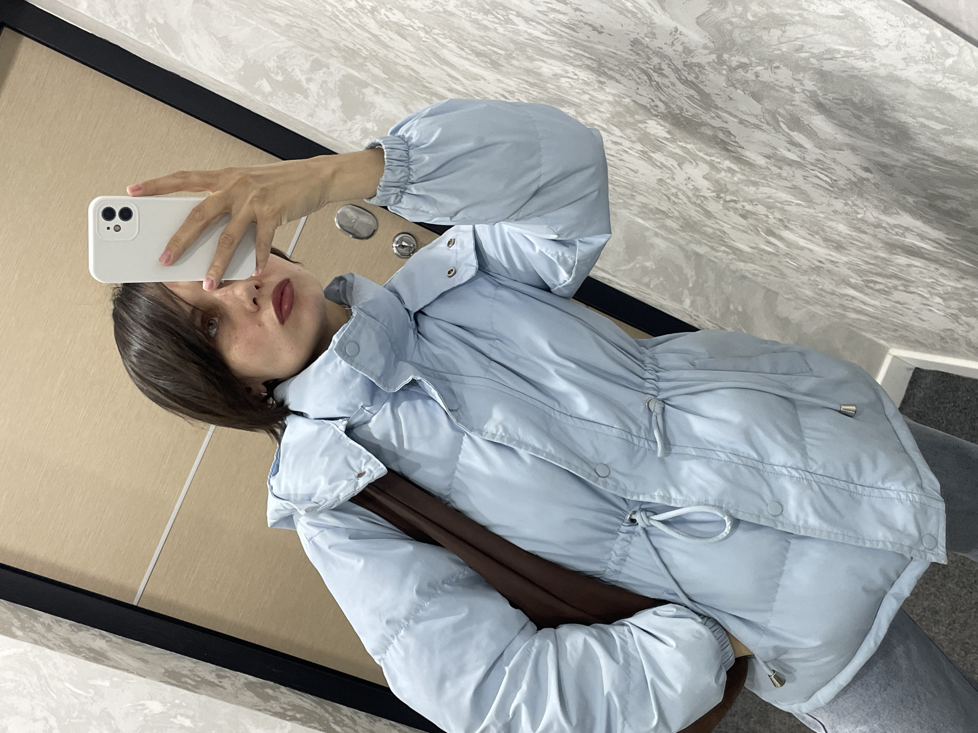 A person with dark hair and fair skin is taking a selfie in what appears to be an indoor setting. The person is holding a white smartphone in their right hand, with the camera facing towards their face. The background includes a combination of wooden panels and grey marble-like patterns. The person is wearing a light blue puffer jacket with a drawstring around the waist. Part of a brown bag or item of clothing can be seen underneath the jacket. There is no indication of Izhevsk, Russia in the scene.