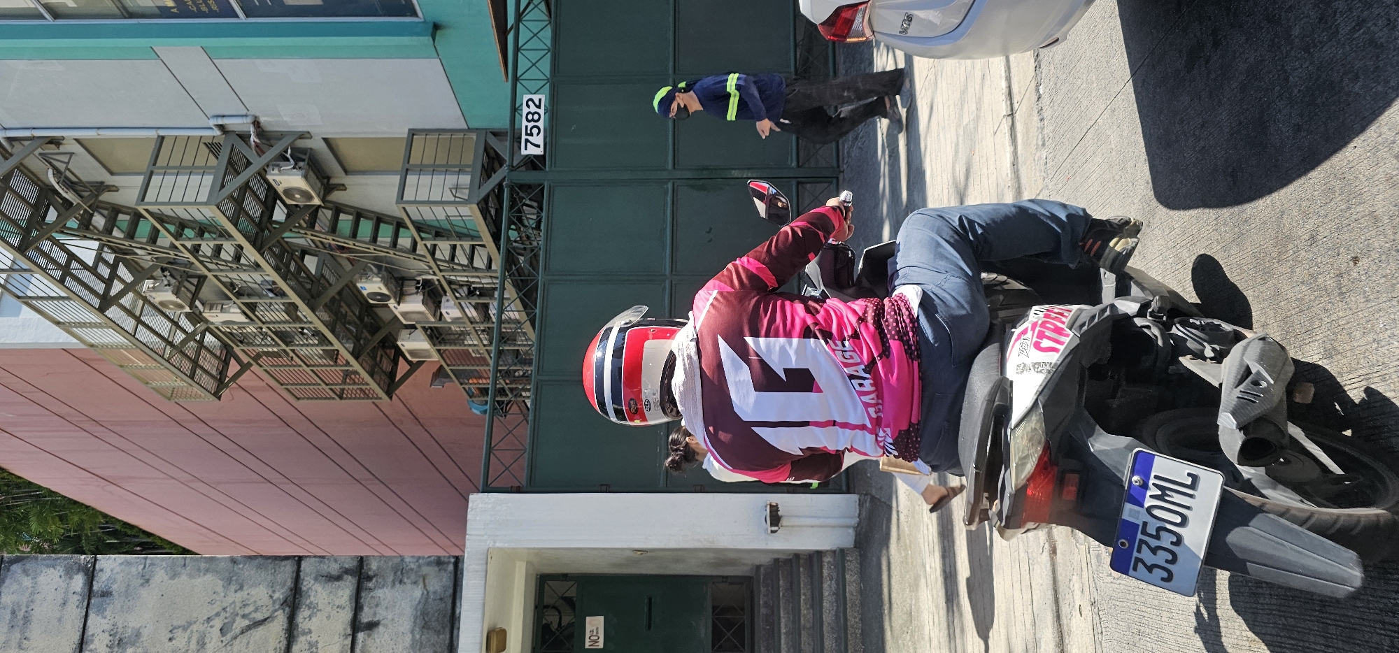 The image captures a street scene in Makati, Philippines, under bright sunlight. In the foreground, a person wearing a red and white helmet and a pink and maroon long-sleeved shirt with a large white "16" graphic and "GARAGE" text is seated on a scooter. The scooter has a license plate with the characters "3350ML". The rider is wearing blue jeans. To the right, a person in a navy blue shirt with lime green stripes and a green bandana is walking, wearing a black face mask. Behind them, a large, dark green metal gate is visible, with the number "7582" displayed on it. To the left, a building with a red facade and horizontal lines is prominent, with an exterior metal staircase attached to a lighter-colored building section. Air conditioning units are mounted on the building. A white building with a green entrance door and a "NO ID" sign is also visible to the left. The ground is paved with concrete. In the far right, the rear of a white car is partially visible. Lush green foliage is seen at the top left of the image.