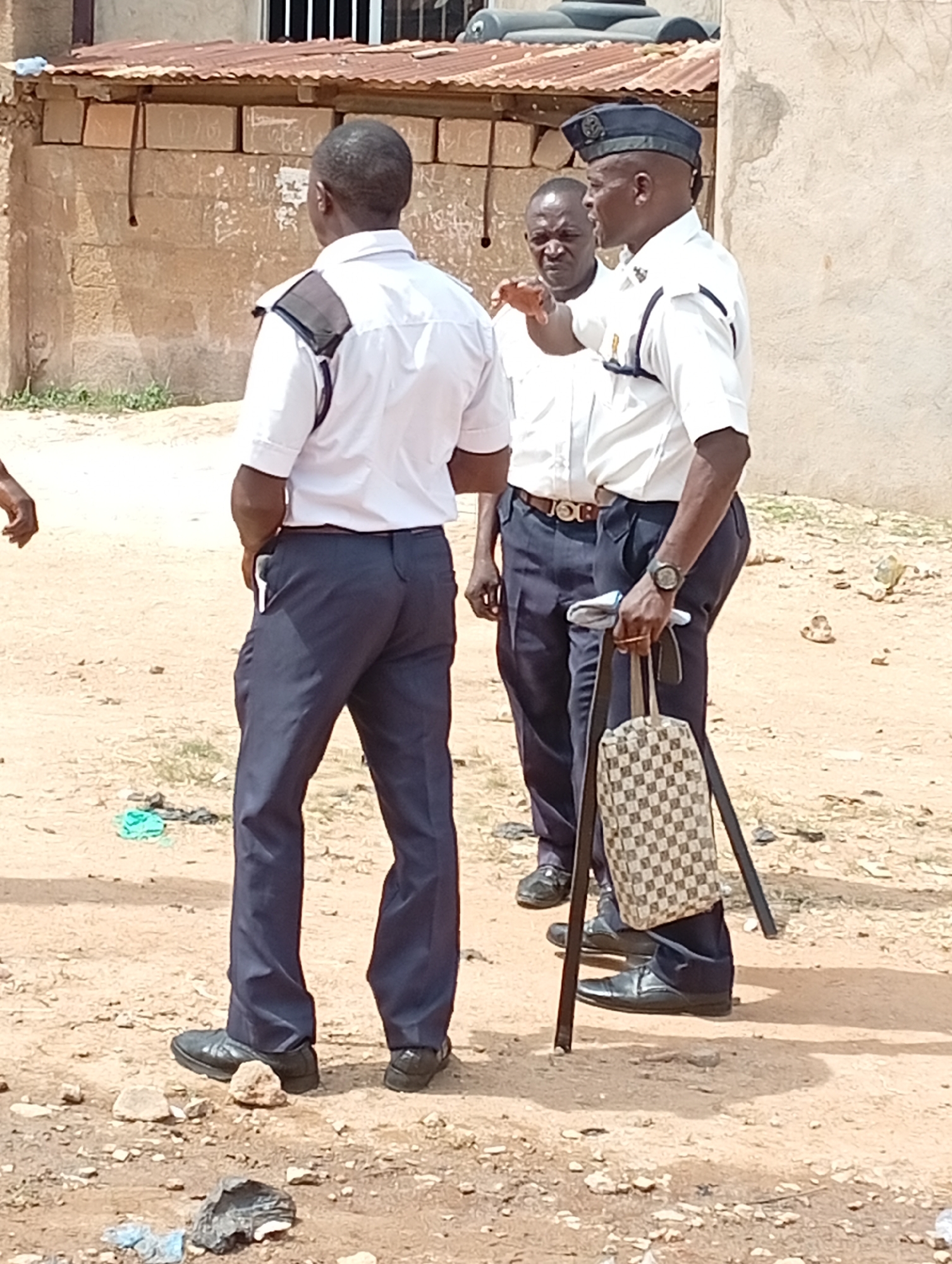 Three men stand outdoors on bare ground. The men are all wearing dark pants and white short-sleeved shirts. The man on the right wears a dark blue cap, carries a checkered tote bag, and has a walking stick. The man in the center is looking at the man on the right and has his hand up. The man on the left has his back to the camera. Behind them is a block building with a corrugated metal roof. There is trash on the ground.