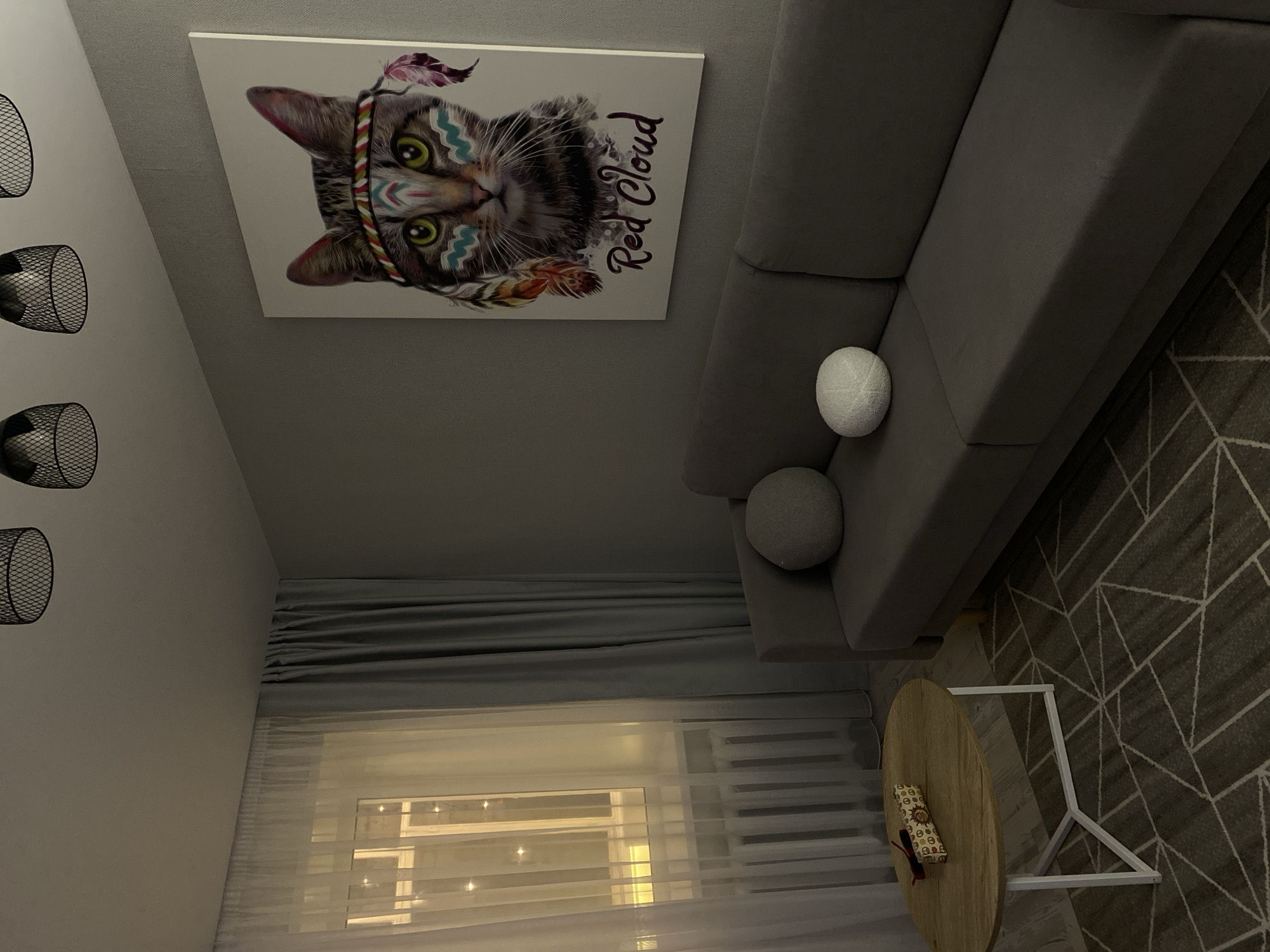 The image depicts a living room scene, likely in an apartment in Votkinsk, Russia. A gray sofa is positioned against a gray wall. On the wall, a large framed canvas art depicts a cat with face paint and feathers, with the text "Red Cloud" below it. Two decorative pillows, one round and gray and the other spherical and white, rest on the sofa. To the left of the sofa, a round wooden coffee table with white metal legs sits on a patterned rug. On the coffee table, a rectangular object with red accents rests. A window with sheer white curtains and darker gray curtains beside it is visible on the left, with string lights illuminated outside. A radiator is located beneath the window. Three black industrial-style pendant lights are suspended from the ceiling. The lighting in the room is dim, with the main light source appearing to be from the window area.