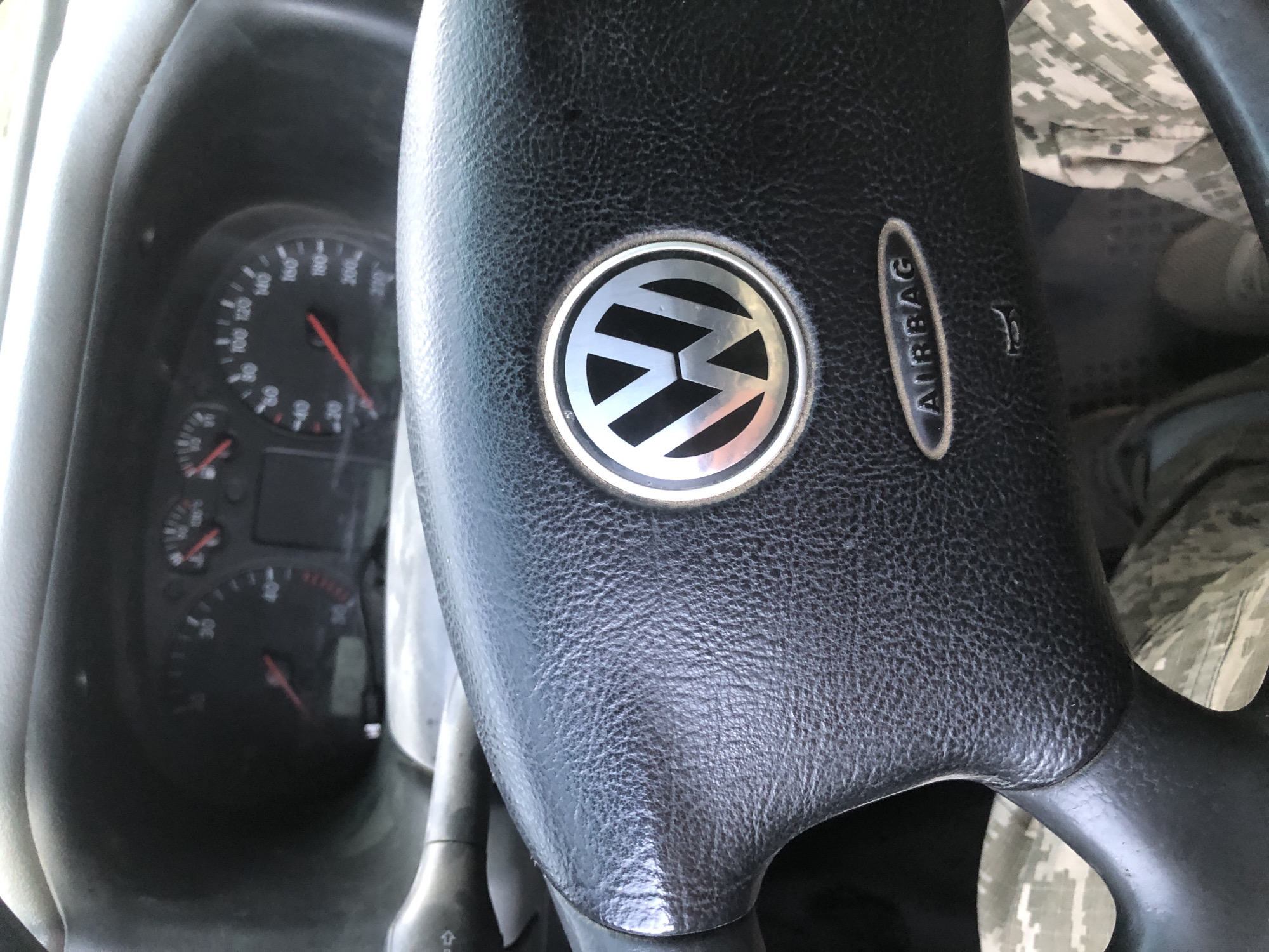 The image is a close-up, eye-level shot of a car's steering wheel and dashboard. The steering wheel is black, textured leather, with the Volkswagen logo prominently displayed in the center. Below the logo, the word "AIRBAG" is embossed in an oval. To the left of the steering wheel, a turn signal lever is partially visible. The dashboard behind the steering wheel shows a speedometer and tachometer, with the needles indicating a speed between 20 and 40 (likely km/h or mph). The background is blurred, showing a hint of camouflage fabric, possibly clothing or seat covers. The overall lighting suggests it is daytime. The location is specified as the city of Mykhalcha, Ukraine.