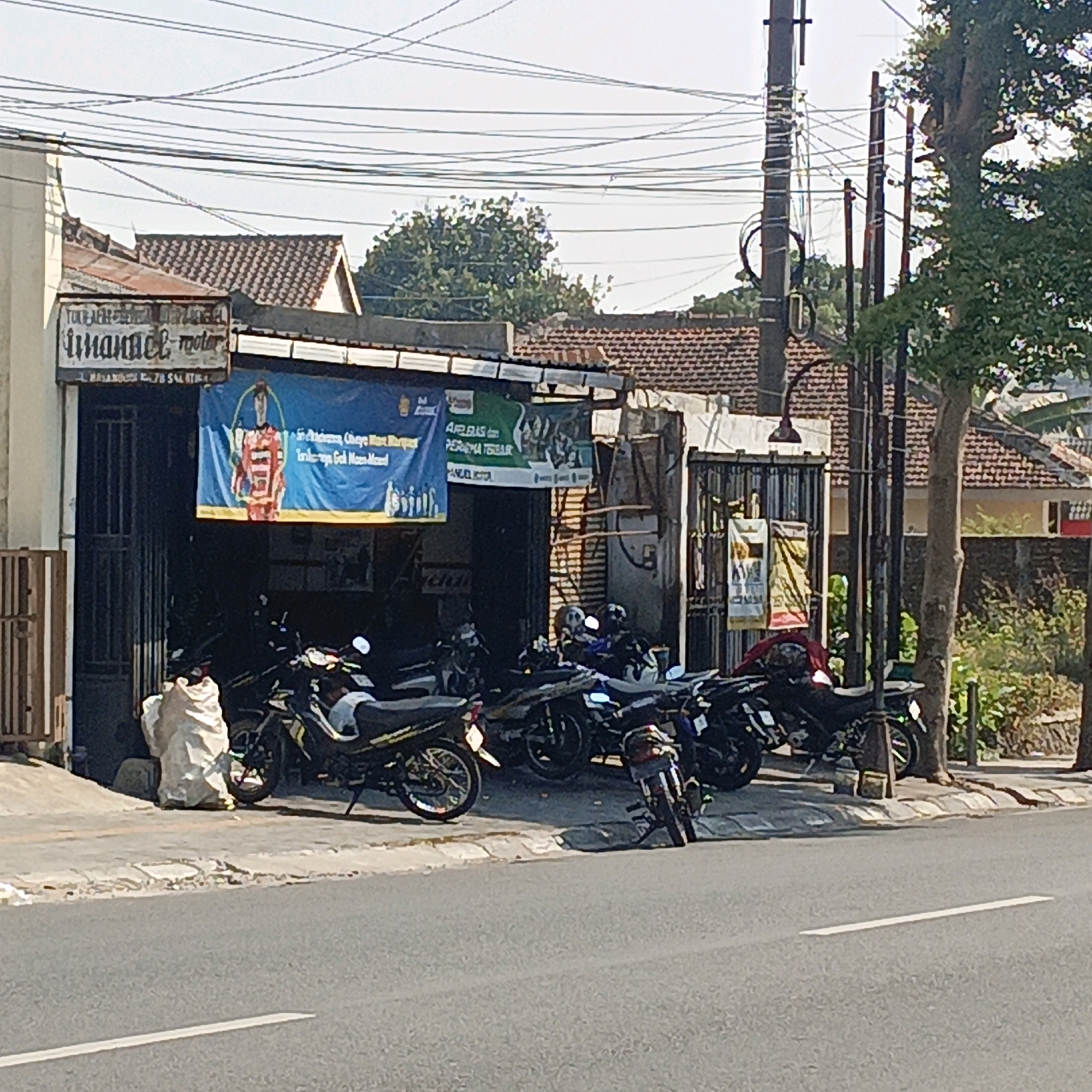 This is a street-level, eye-level, medium shot of a motorcycle repair shop on a sunny day in Salatiga, Indonesia. The shop, named "LINAMAX MOTOR", features a blue banner with white text and an illustration of a person in a red outfit. Several motorcycles are parked in front of and inside the open shop. A large, light-colored sack is placed on the ground near the entrance. The building has a metal roll-up door and is flanked by a wooden fence-like structure on the left. The shop is situated on a paved street with a sidewalk. Electrical poles and wires are visible in the background, along with the roofs of nearby buildings and trees. The overall scene depicts a typical roadside business in an urban Indonesian setting.