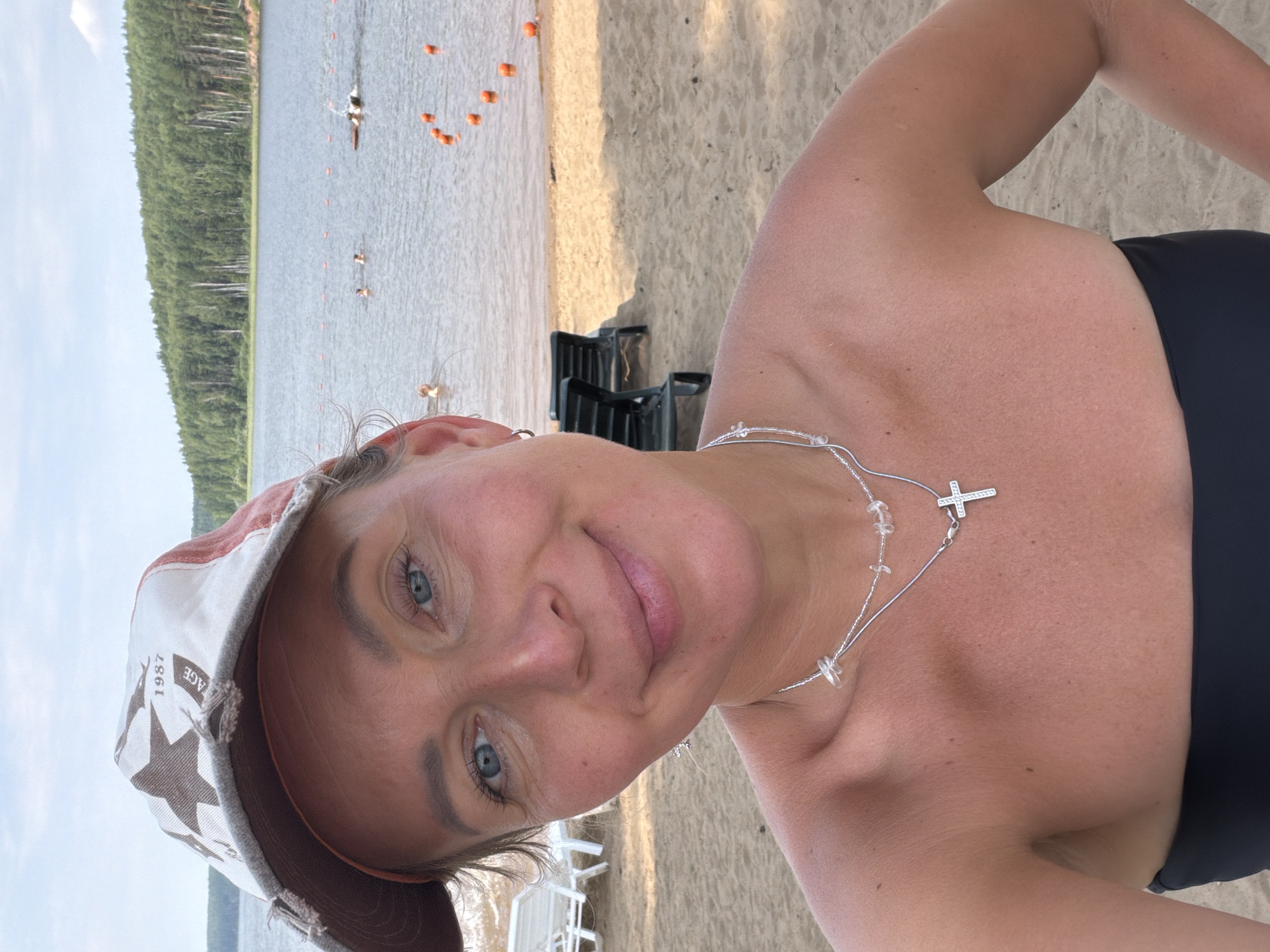 In a selfie taken on a sandy beach, a woman wearing a baseball cap and a black strapless top smiles at the camera. She has blue eyes and light skin, and is wearing a necklace with a cross pendant. In the background, a lake is visible with people swimming and a line of orange buoys. Trees line the far shore of the lake, suggesting a natural or park-like setting. Beach chairs are set up on the sand behind her, indicating a recreational area. The location is the Kama River beach in Chaykovsky, Russia.
