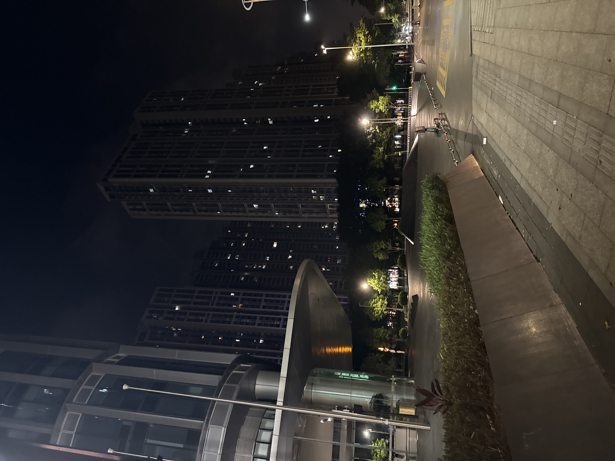 This is a nighttime photograph taken in Haizhu District, China. The scene features several tall residential buildings with many illuminated windows, suggesting that it is evening or night. In the foreground, there is a modern building with glass facades and metallic accents, partially obscuring the view of the residential towers. To the right, a paved walkway is visible, bordered by a neatly trimmed hedge. Streetlights illuminate the area, casting a warm glow on the wet pavement, indicating recent rain. Lush green trees are interspersed between the buildings and along the walkway, their leaves reflecting the artificial light. There are no people visible in the frame, creating a sense of quiet and stillness. The overall mood is serene and urban, highlighting the nighttime cityscape. No visible text is present in the image.