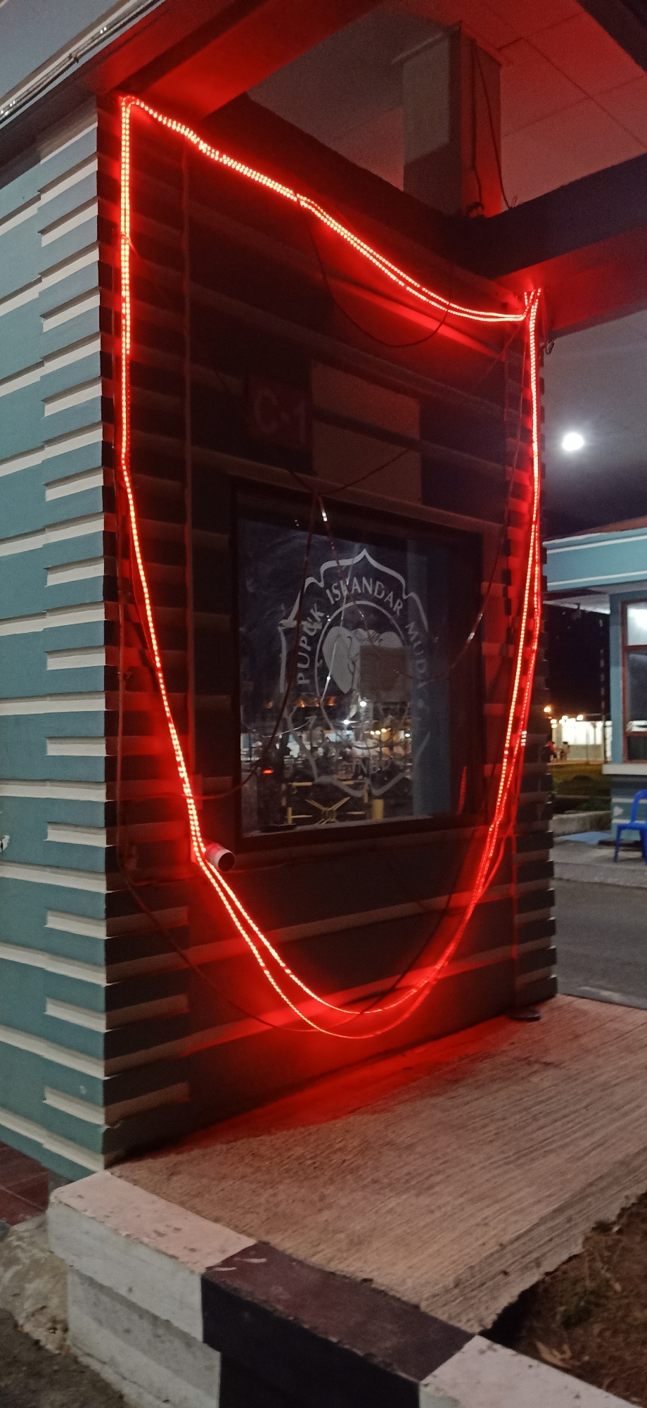The image shows the exterior of a building at night, likely a guardhouse or entrance to a facility. The building has a segmented, light blue facade and a darker, wood-paneled section. A prominent feature is a string of bright red LED lights outlining a U-shaped or horseshoe shape around the darker section of the building, casting a warm glow on the surrounding concrete and facade.
The darker section features a rectangular window, through which a logo or emblem is visible. The emblem is circular and appears to contain an elephant motif with text around it. The text is partially obscured but includes "PUPUK ISKANDAR MUDA". Above the window, a sign with "C-1" is visible. Inside the window, interior lighting and faint reflections suggest activity within the building.
The scene appears to be outdoors, with a concrete ground surface in the foreground marked with black and white stripes along the edge. To the right, a blue plastic chair is visible, and in the background, other buildings and lights are faintly illuminated, suggesting an urban or developed area. The overall mood is illuminated and possibly welcoming due to the bright lighting. The time of day is clearly night. There are no people clearly visible in the foreground, but faint figures can be seen through the window. The weather appears calm, given the clear visibility and lack of atmospheric disturbance.