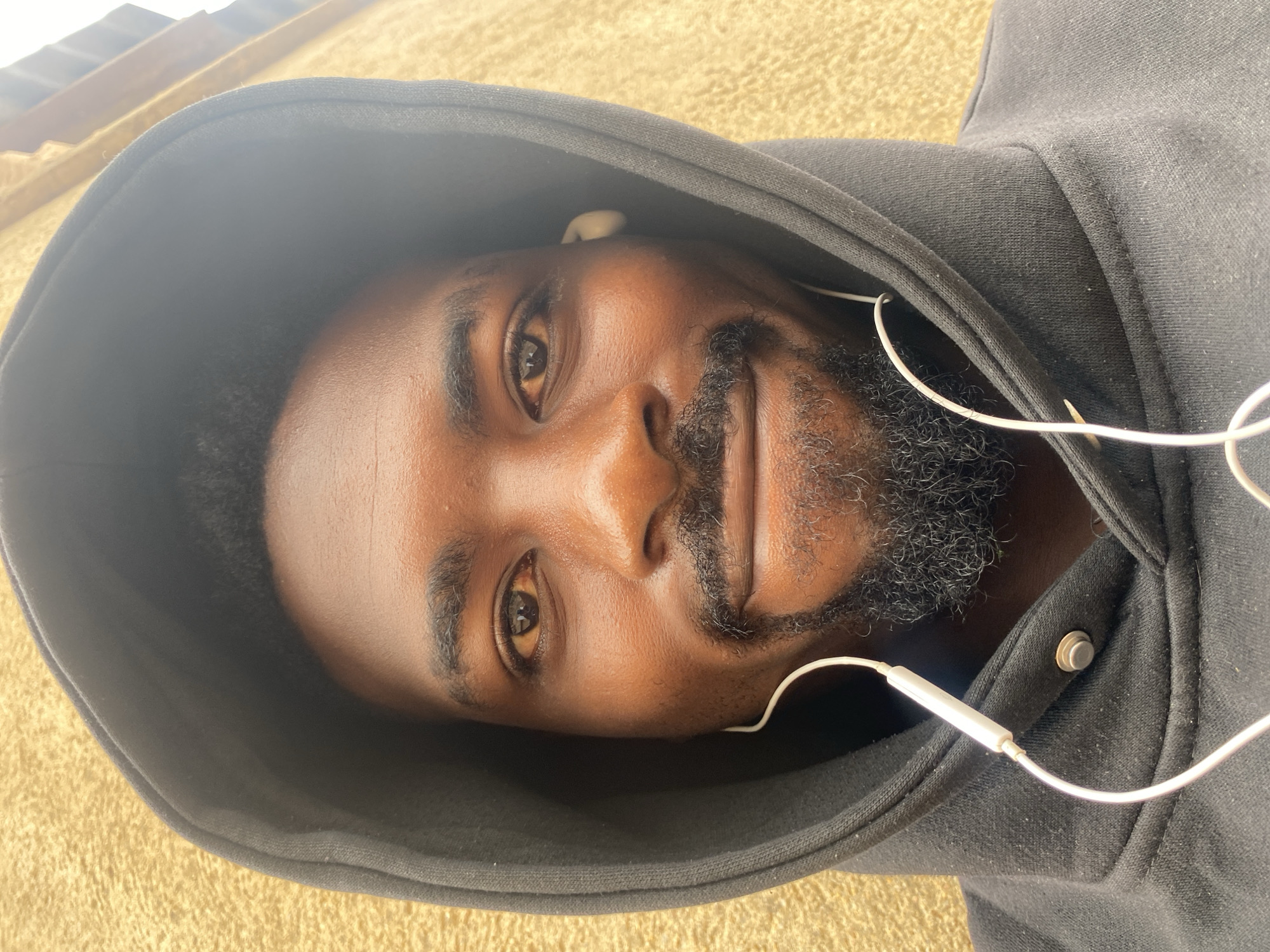 A man with dark skin is wearing a black hooded sweatshirt with the hood up and earbuds in his ears. He has short black hair and a beard. He is looking directly at the camera. The background is a beige or light brown color. The location is not discernible from the image.