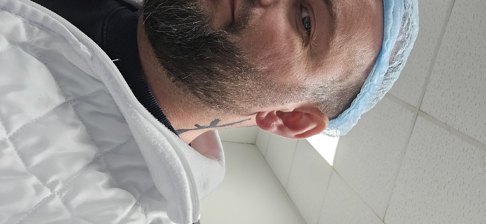 A close-up, tilted shot shows a person's face and upper body. They are wearing a light blue disposable hairnet. The person has a beard and a tattoo on their neck. They are wearing a white quilted jacket or coat. The background features tiled walls and a ceiling. The location cannot be determined from the image.