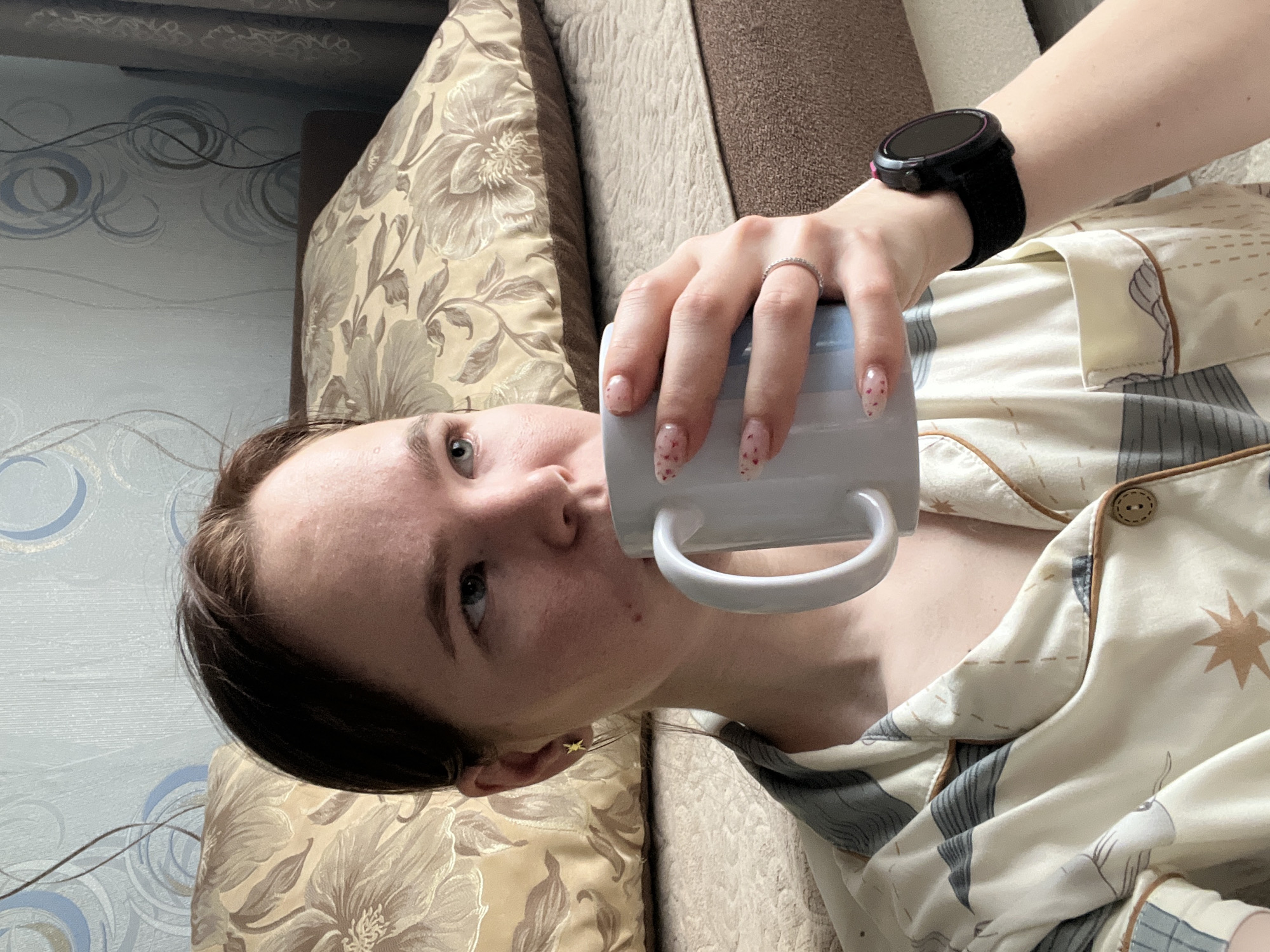 A woman is holding a light blue mug to her lips, possibly drinking from it. She is wearing a ring on her finger, a smartwatch on her wrist, and what appears to be pajamas. The environment includes a floral-patterned pillow and what seems to be a couch or bed. The location is Chaikovsky, Russia.