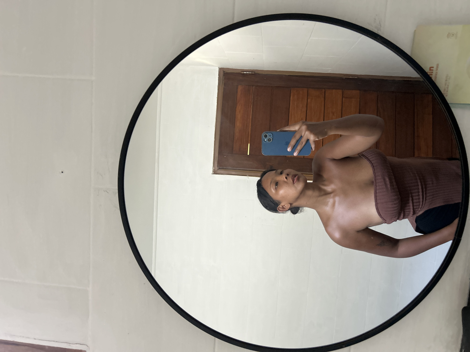 A woman is taking a mirror selfie in a bathroom. She is holding a blue smartphone in her right hand, capturing her reflection. She is wearing a brown strapless top and black bottoms. The bathroom features white tiled walls and a wooden door. The mirror is round with a black frame. A yellow package with some text on it is visible on the counter to the right. The scene appears to be in Kuta, Indonesia, based on the contextual information provided.