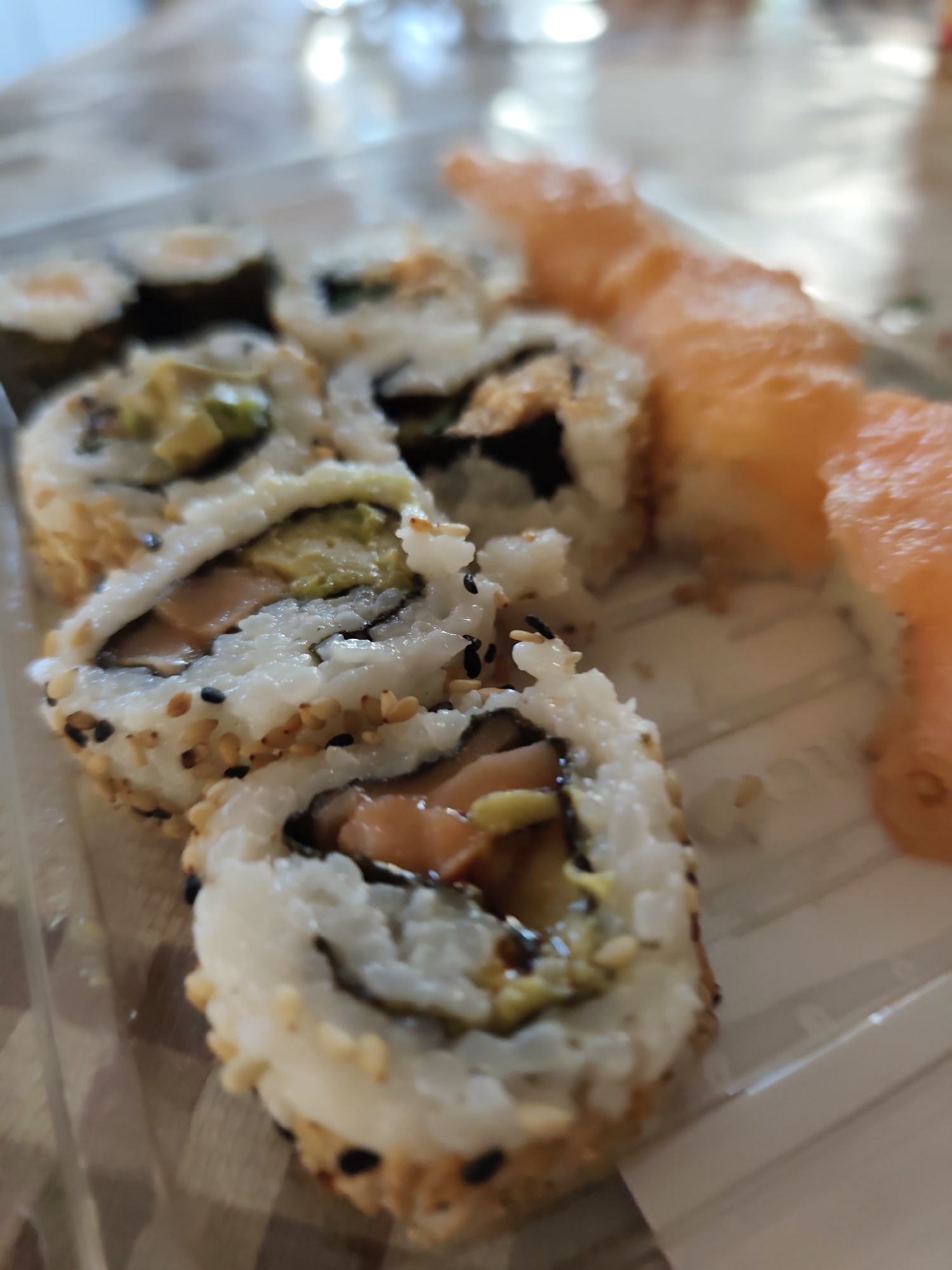 A close-up, shallow depth of field, indoor shot shows a clear plastic container filled with several pieces of sushi and sashimi. The sushi rolls are made with white rice, seaweed, and appear to contain avocado and salmon. They are coated with sesame seeds, some white and some black. The sashimi is a bright orange salmon, sliced thinly and arranged on the side. The background is blurred, suggesting an indoor setting, possibly a table or counter. The lighting is bright, with reflections visible on the surface of the container and the food. There are no people visible in the image. The overall scene is clean and appetizing, highlighting the freshness of the seafood. Without further information, the specific location in Castlebar, Ireland, cannot be determined.