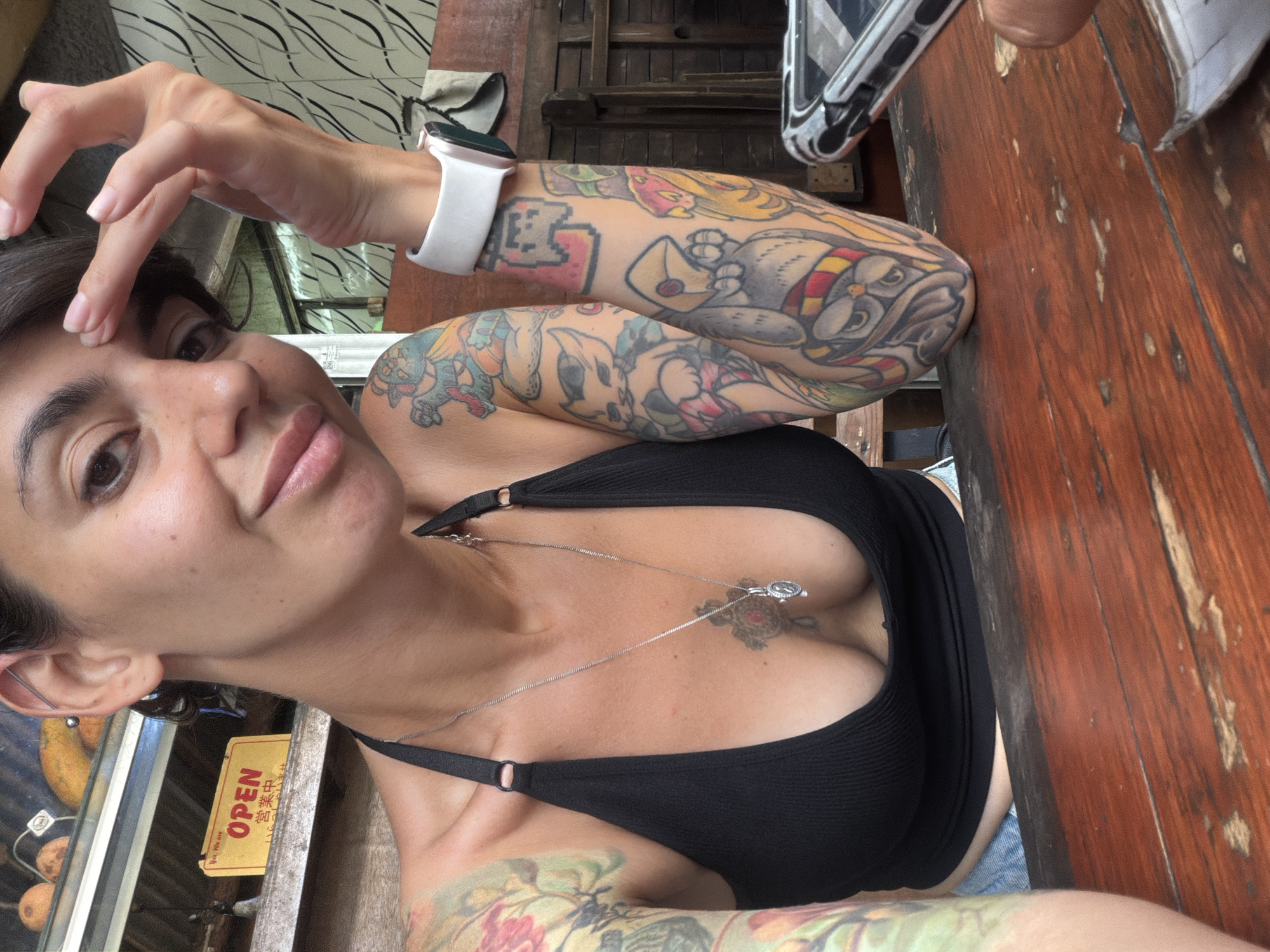 A woman with multiple colorful tattoos on her arms and a chest tattoo is captured in a close-up, high-angle shot. She is seated at a rustic wooden table, with a portion of a smartphone visible on the right side of the frame. The woman is wearing a black halter top and a silver necklace with a pendant. She is touching her forehead with her right hand. In the background, a shelf displays some fruits, and a yellow sign with "OPEN" in English and Japanese characters is visible. The overall setting appears to be a casual eatery in Kuta Selatan, Indonesia.