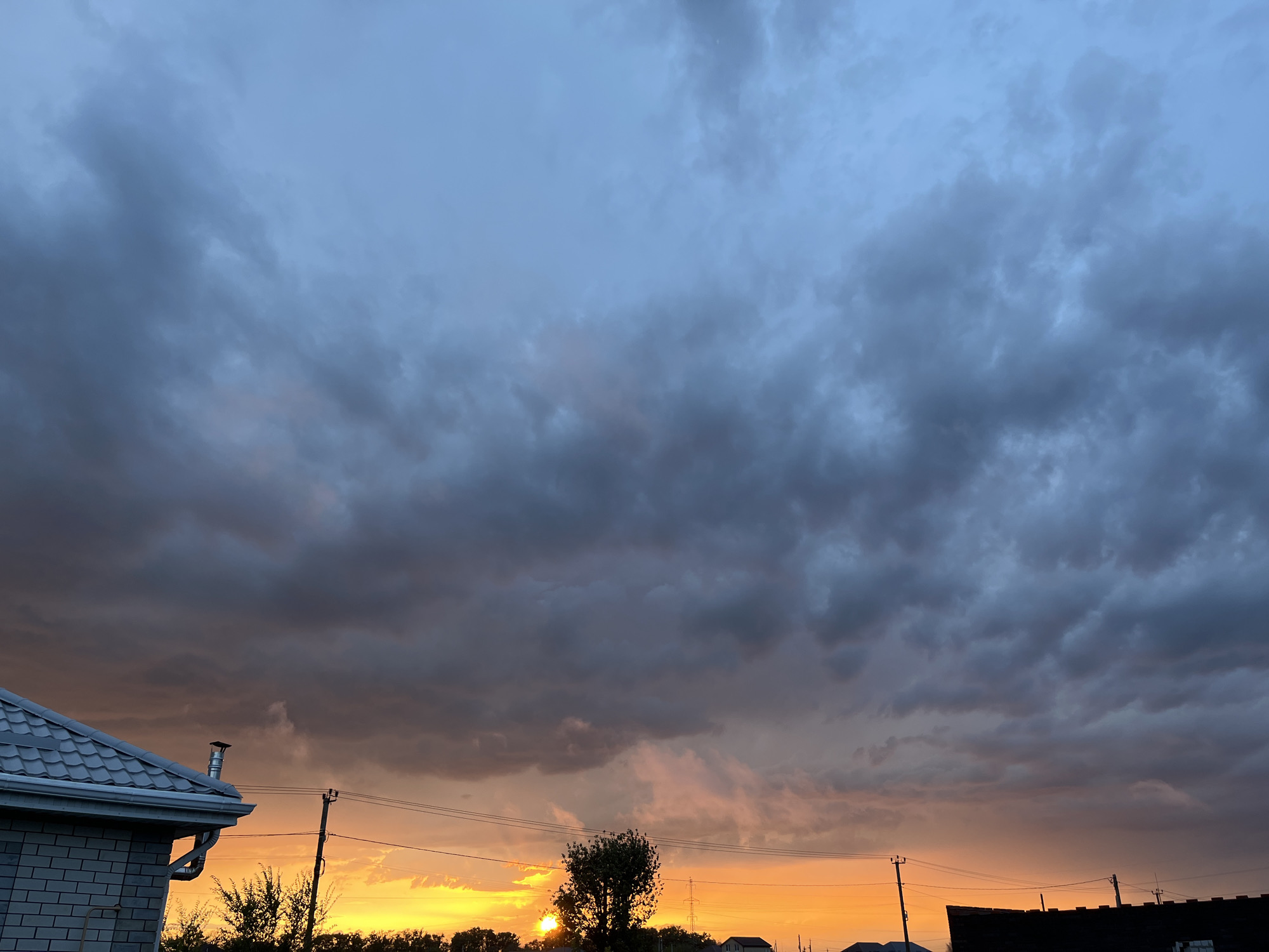 The image depicts a sunset with dramatic clouds over a suburban or rural landscape. The sky is dominated by dark, ominous clouds that transition to hues of orange and yellow near the horizon, indicating the setting sun. Silhouetted against this colorful sky are trees, utility poles, and the rooftops of several buildings, including a house with a tiled roof and a brick wall on the left side of the frame. The overall atmosphere is moody and atmospheric, characteristic of a stormy or overcast sunset. The scene is set in Taganrog City District, Russia.
