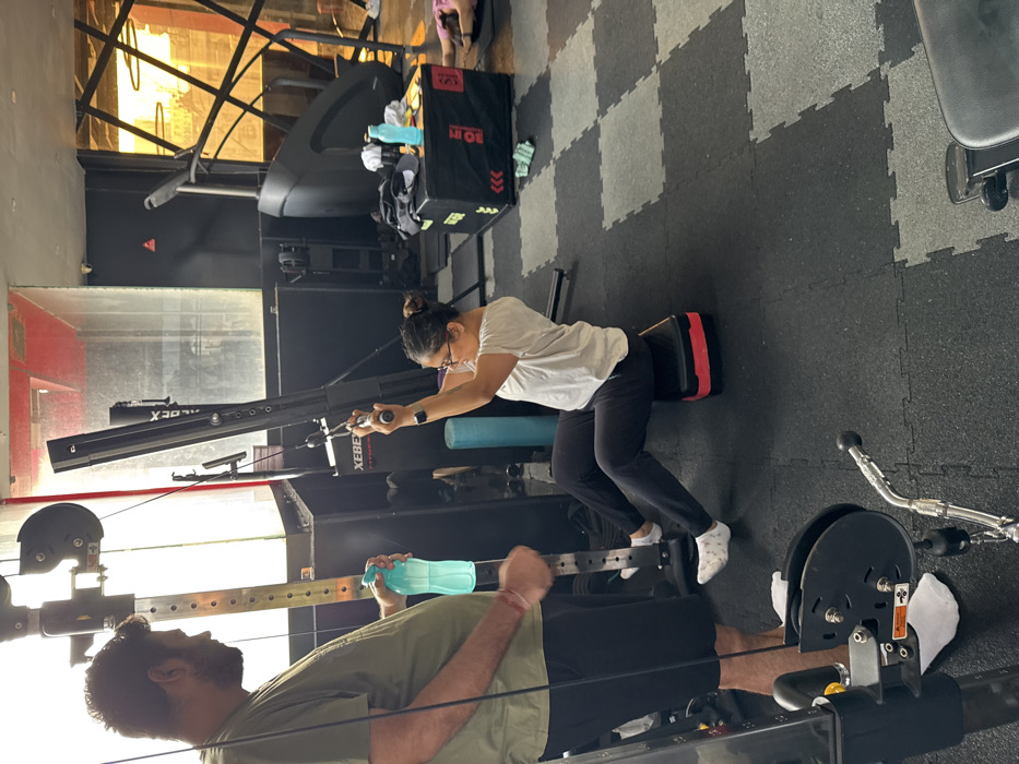 In a gym setting, a woman in a white t-shirt and black pants kneels on a pad, pulling a cable machine towards her. Nearby, a man in a green shirt and black shorts is positioned on a rowing machine. Gym equipment like a treadmill and weights are in the background, and the floor is covered in interlocking black and gray tiles. Natural light streams into the space.