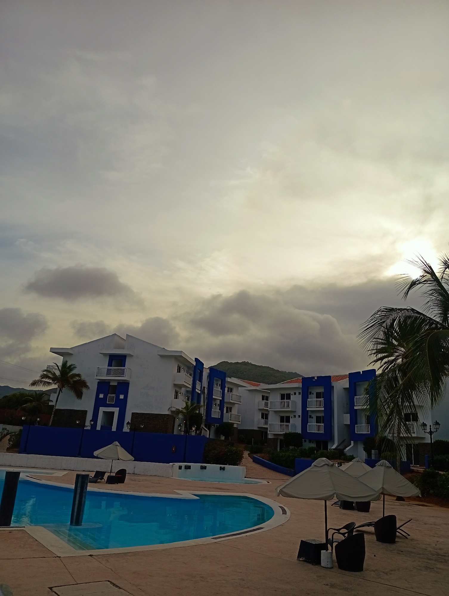 The image depicts a pool area in Playa El Agua, Venezuela, with a multi-story building complex featuring white walls and blue accents. The building is set against a cloudy sky, with hints of mountains in the background. A palm tree is visible on the right side of the frame. In the foreground, there is a swimming pool with clear blue water. Several patio umbrellas and chairs are positioned around the pool deck. The lighting suggests either early morning or late afternoon due to the overcast sky.