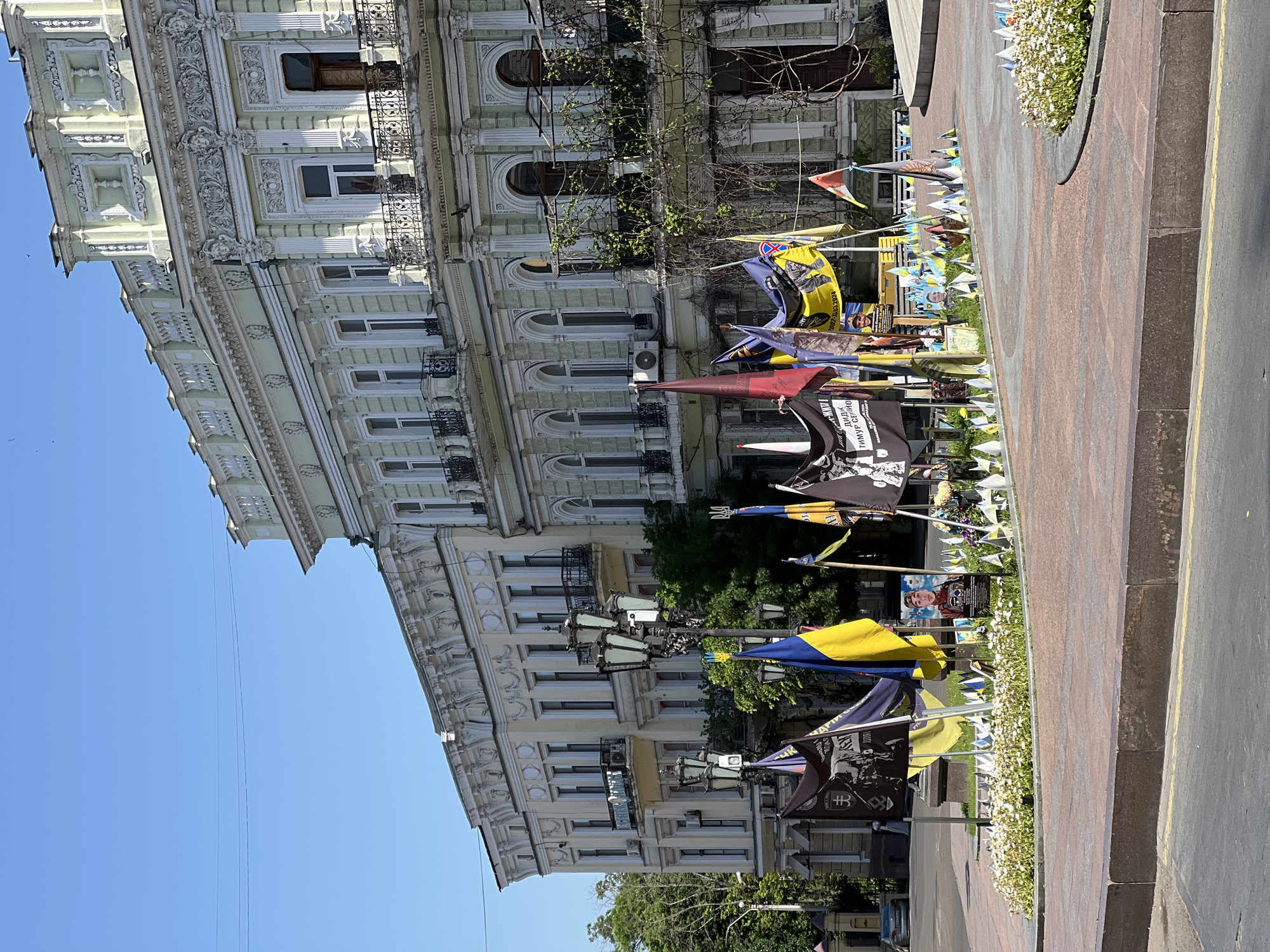The image shows a street scene in Odesa, Ukraine, likely taken in the daytime. In the background is a building with ornate architectural details, including decorative moldings, windows, and balconies. The facade is light-colored.
In the foreground, a row of flags and banners lines the street, possibly placed along a sidewalk or pedestrian area. Some of the flags display the Ukrainian national colors, blue and yellow. Other flags and banners display what appears to be commemorative images and symbols.
A raised curb or barrier separates the flags and banners from the street. Flowers grow between the sidewalk and the street. The sky is clear blue, visible in the upper left corner.