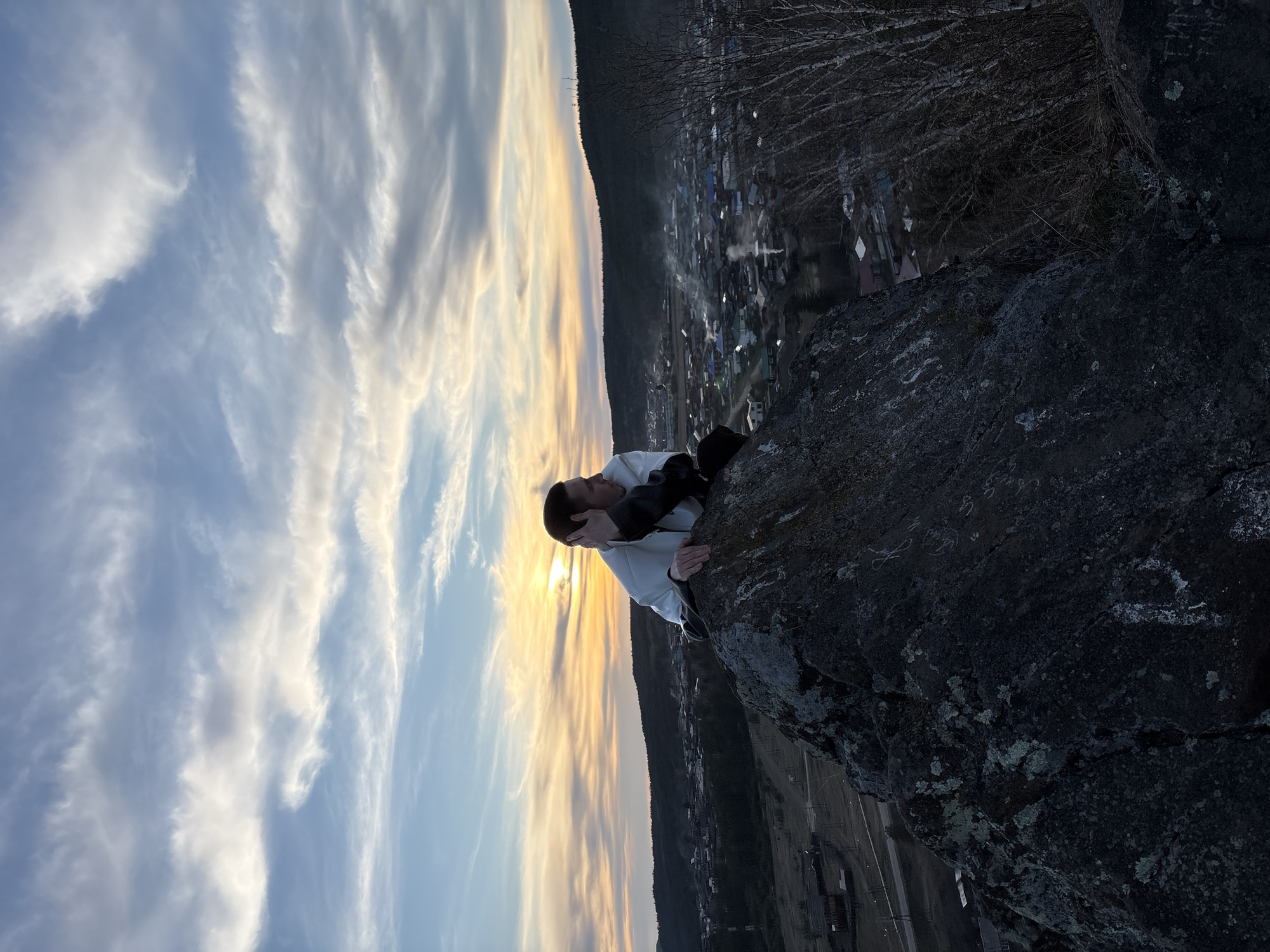 A person is sitting on a large, dark-colored rock at the edge of a cliff. The person is wearing a white hooded sweatshirt and dark pants, with their arms crossed and head resting on their hands. The setting sun is directly behind the person, casting a warm glow on the sky and creating a silhouette effect. In the background, there is a village or town, possibly Невон, Russia, nestled in a valley with a mountain range in the distance. The sky is filled with white and grey clouds.