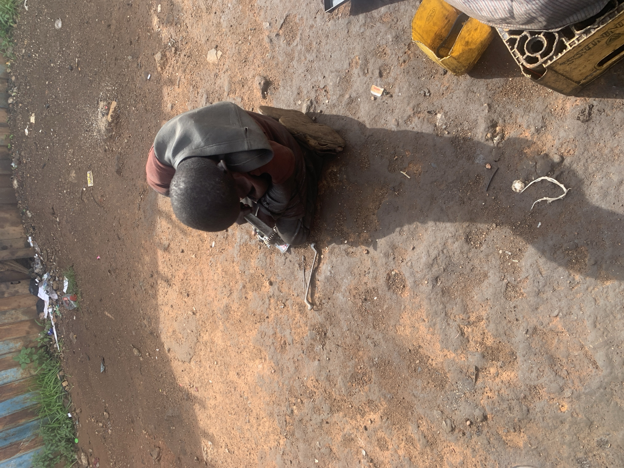 A young boy is seated on the ground in Gura Top, Nigeria, hunched over and seemingly engaged in an activity with small objects. He is wearing a dark gray hooded garment over a reddish-brown shirt and dark pants. His head is bowed, obscuring his face, and his hands are positioned near something on the ground in front of him. The environment appears to be an outdoor area with a dirt and gravel surface. Scattered debris, including pieces of plastic and other small items, are visible on the ground. In the background, a corrugated metal fence stands, with some greenery growing near its base. To the right of the boy, a yellow plastic container and a plastic crate are present. The lighting suggests it is daytime, with shadows cast on the ground.