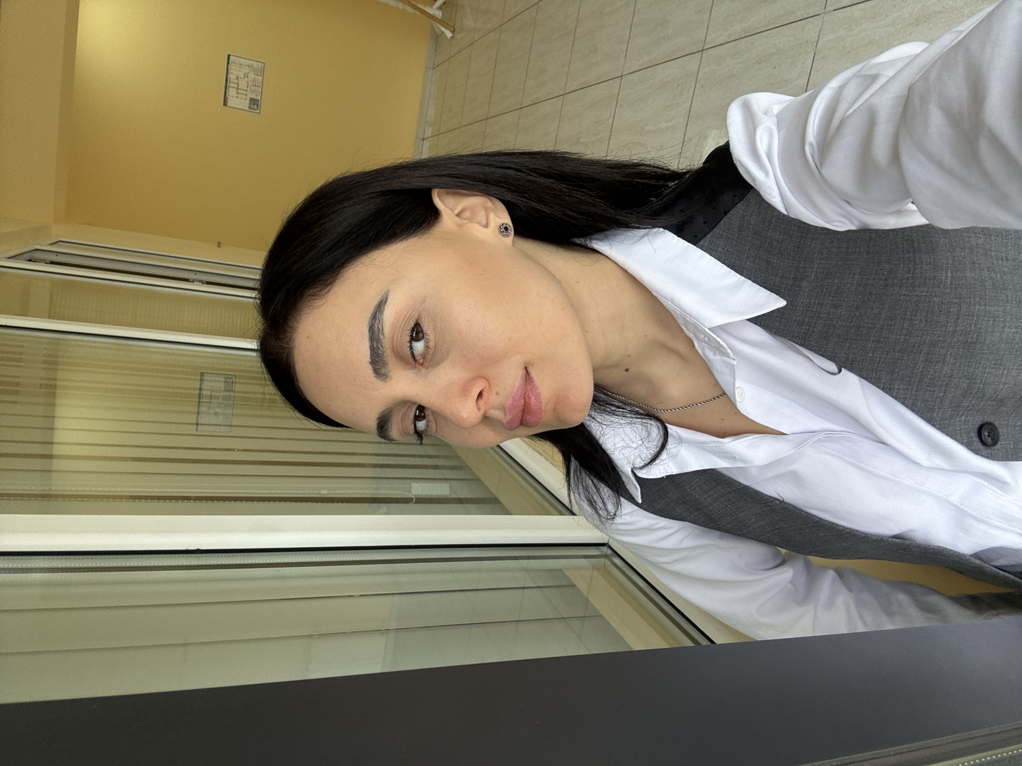 A woman with dark hair is seen in a close-up shot, wearing a white shirt and a gray vest. She is positioned near a window with blinds. The location is likely Tbilisi, Georgia. The background includes a yellow wall with a sign. The foreground shows a black surface.