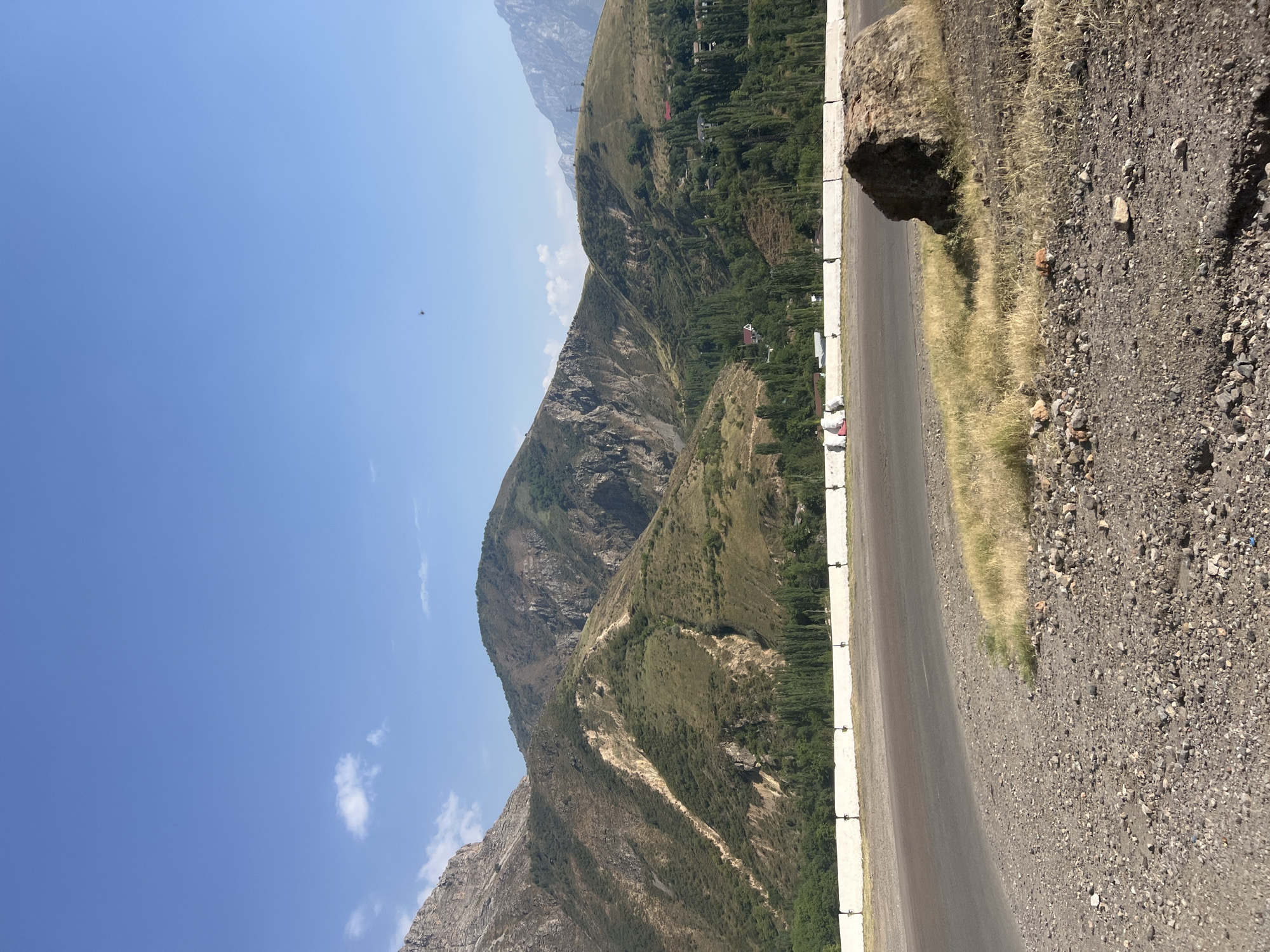 A wide shot captures a majestic mountain landscape under a clear blue sky with sparse white clouds. The foreground features a paved road curving into the distance. To the left of the road, a rocky, gravelly terrain is visible, with patches of dry grass and a large, rough-hewn boulder resting on the grass. A white concrete barrier with metal posts separates the road from a grassy embankment that slopes up towards a densely forested valley. Beyond the trees, the rugged mountains rise, their slopes a mix of green vegetation and exposed rock faces. Several small buildings with red roofs are nestled within the trees, suggesting a small settlement. The overall impression is one of natural beauty and rural tranquility.